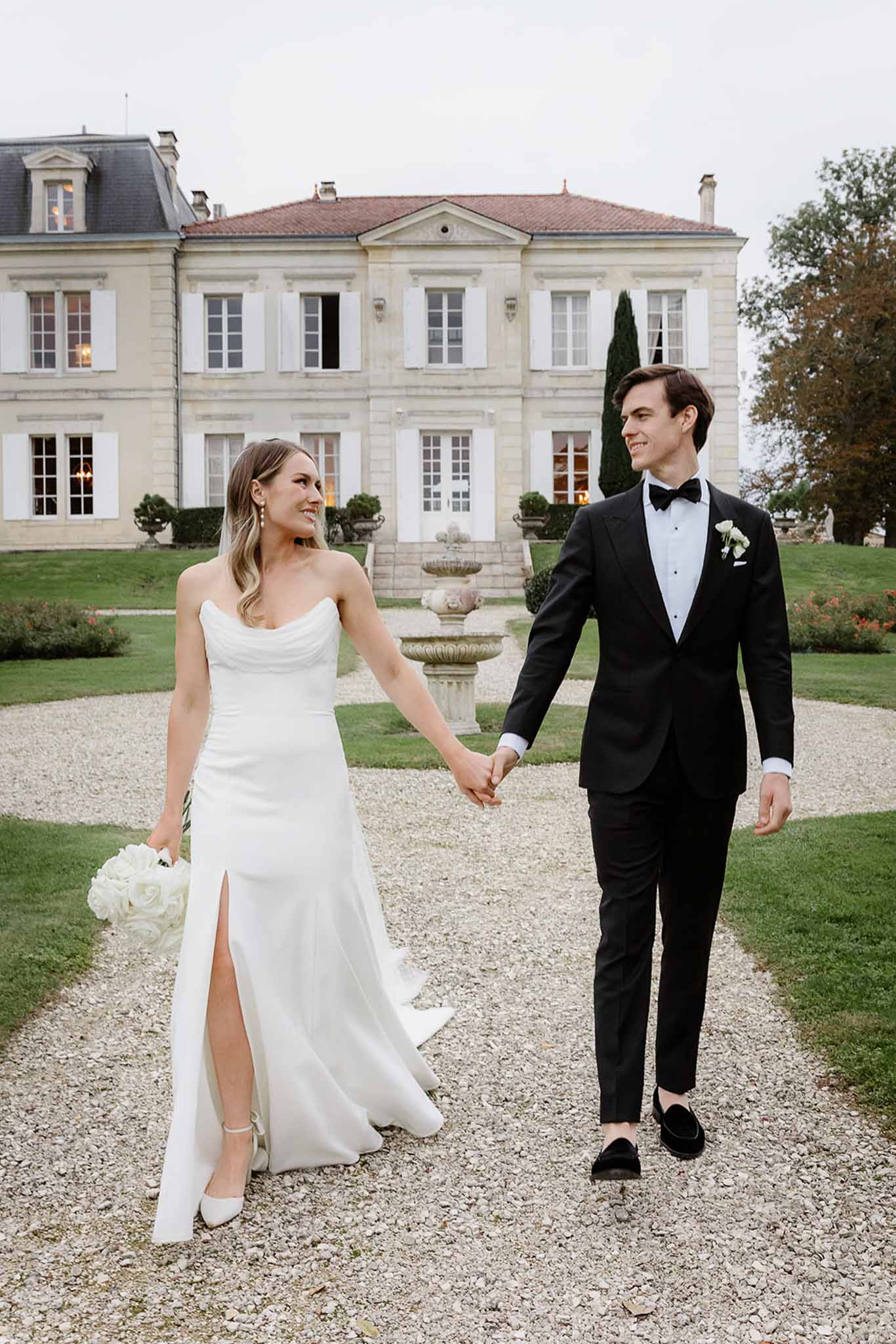 Bride and groom walking hand-in-hand along gravel pathway at French château with neoclassical architecture