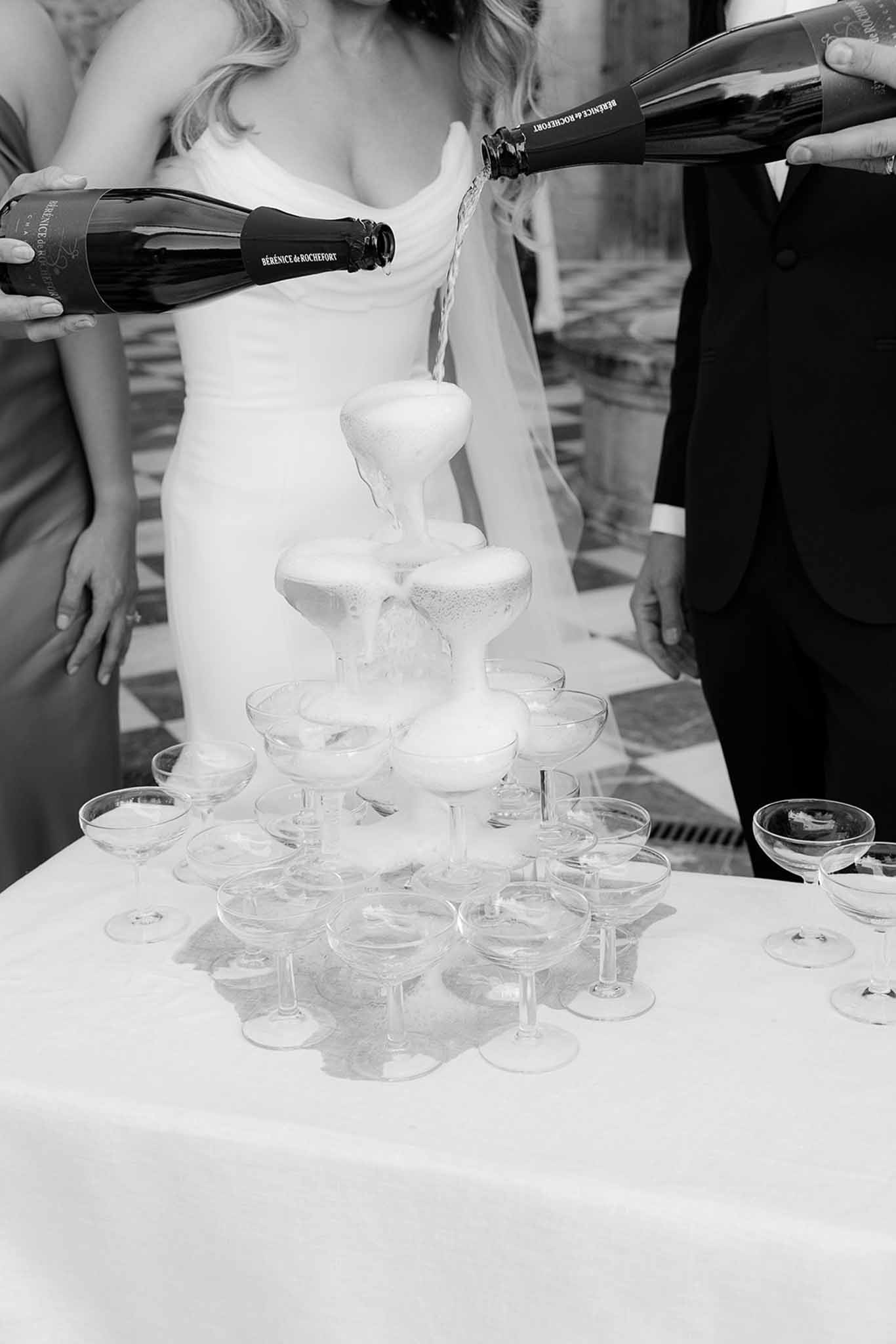 Black and white champagne tower pour with cascade of coupe glasses, bride and groom behind tower