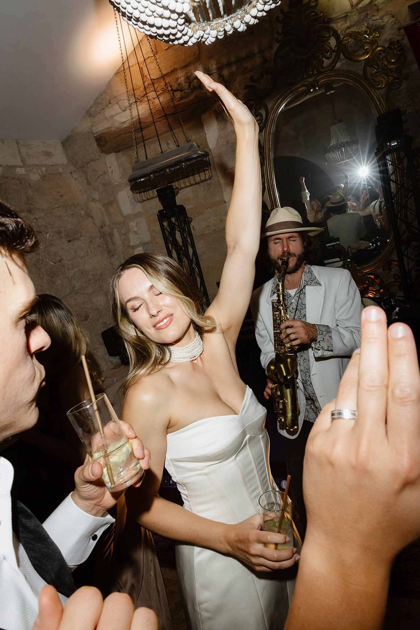 Bride dancing with champagne glass during reception in stone-walled venue with live saxophone music