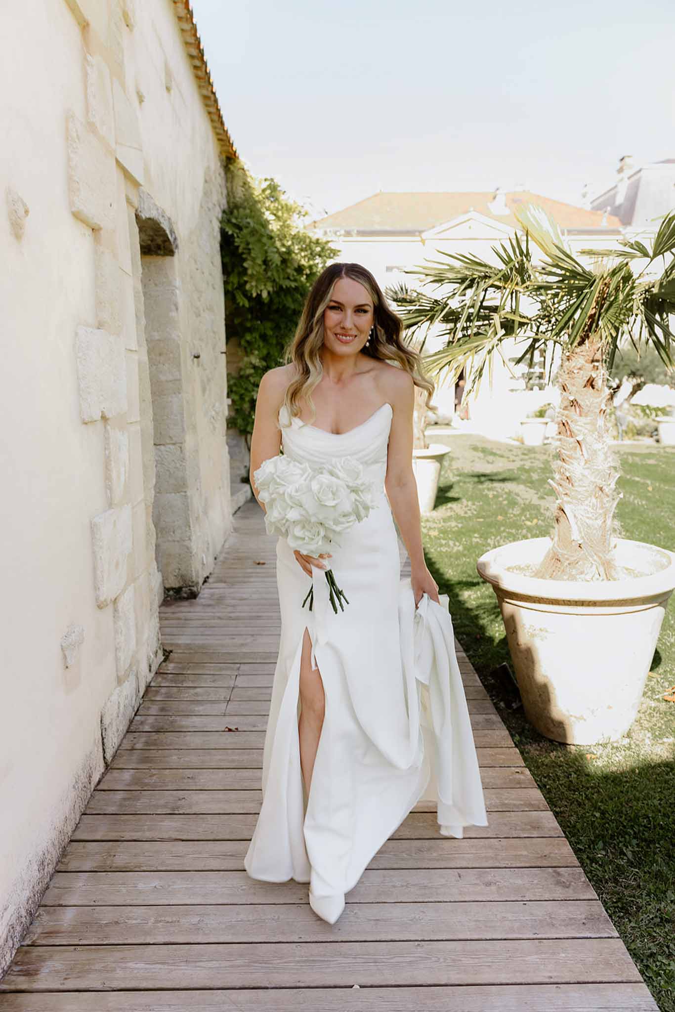 Bride in ivory strapless dress with white rose bouquet on Mediterranean venue boardwalk with palm trees