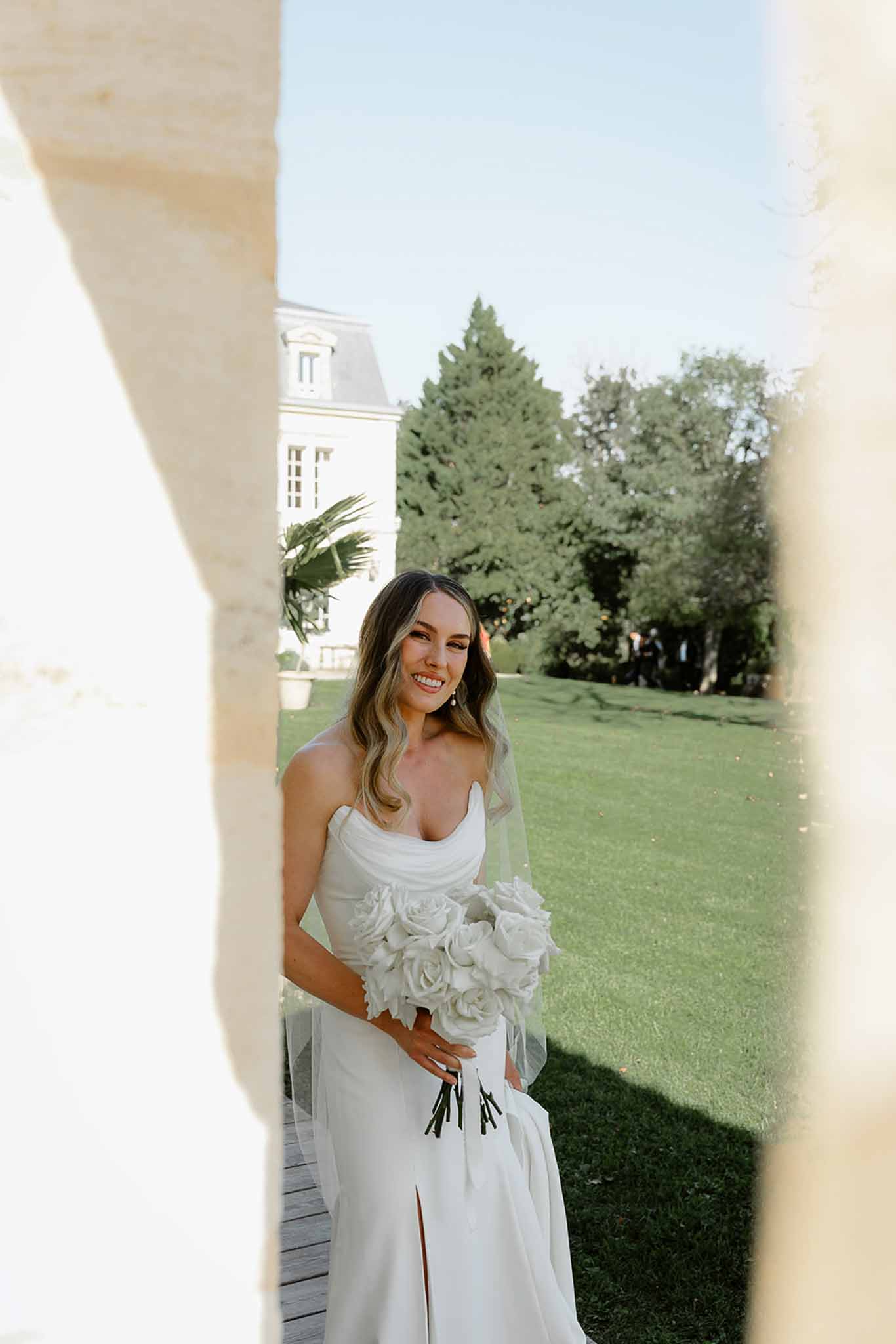 Bride in ivory dress with white rose bouquet posing between cream columns at elegant garden venue