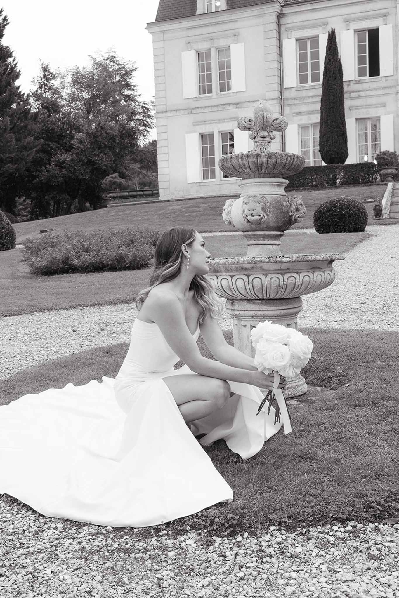 Bride in wedding gown sitting in formal gardens of French estate with neoclassical manor house