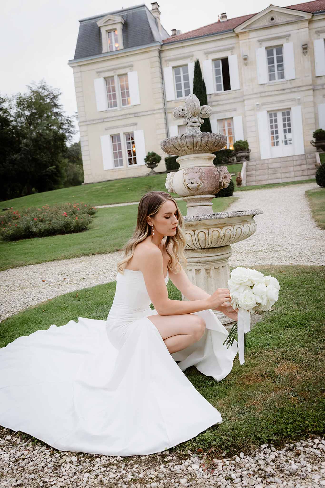 Bride in ivory dress with white bouquet posing by stone fountain at French château courtyard