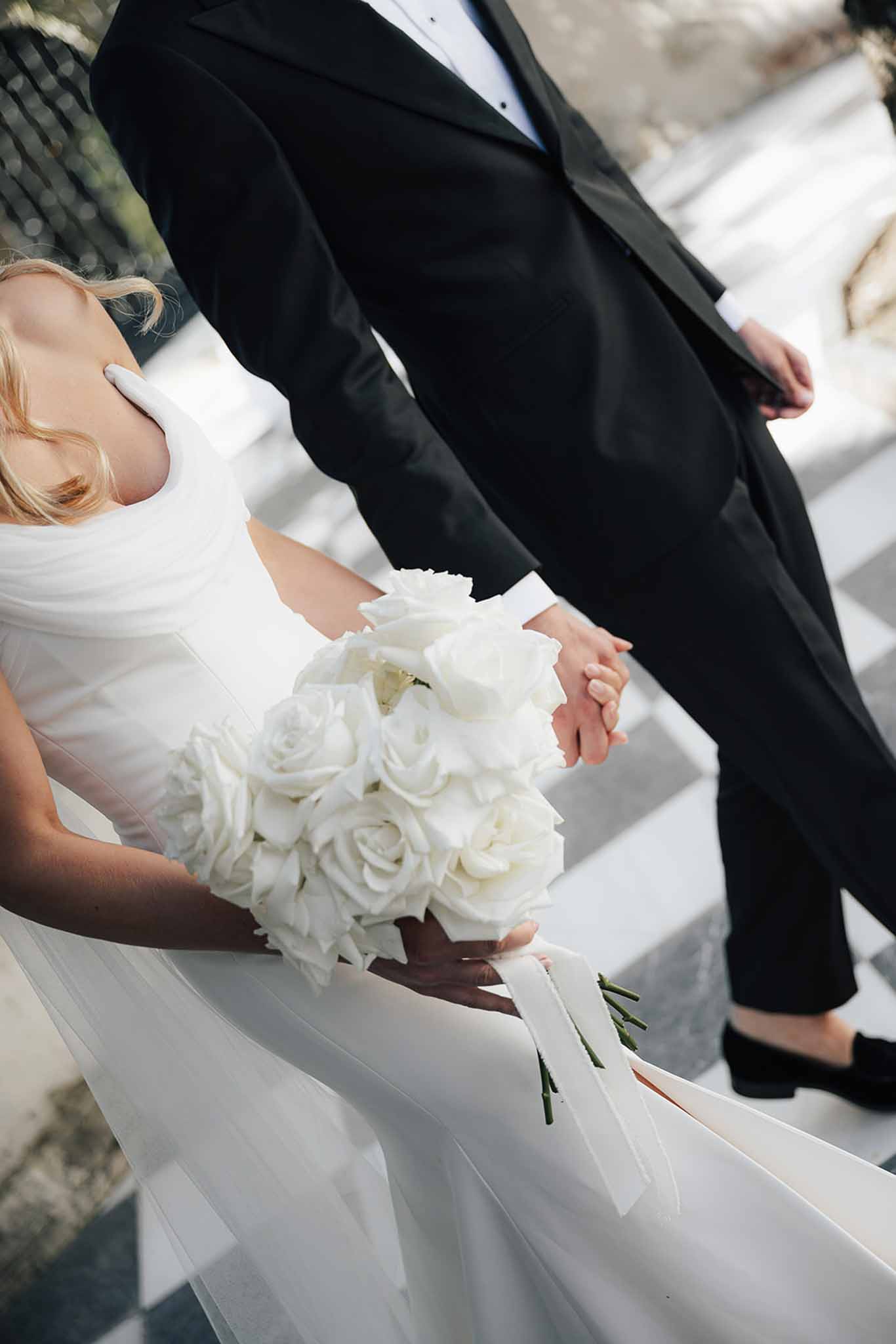 Close-up of bride and groom holding hands, white rose bouquet with ribbons on checkered stone floor