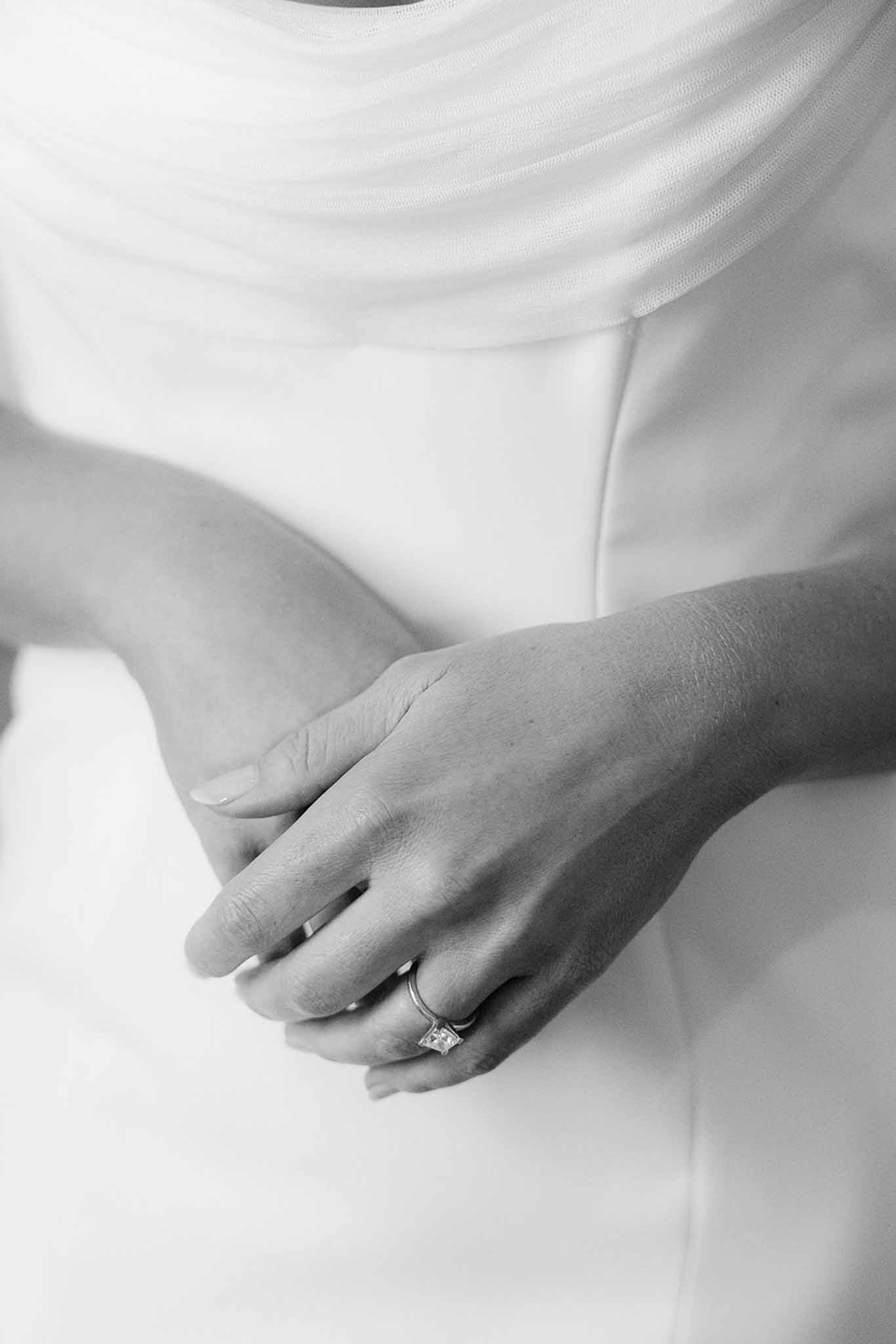Black and white close-up of bride's hands with square-cut solitaire ring over draped tulle bodice