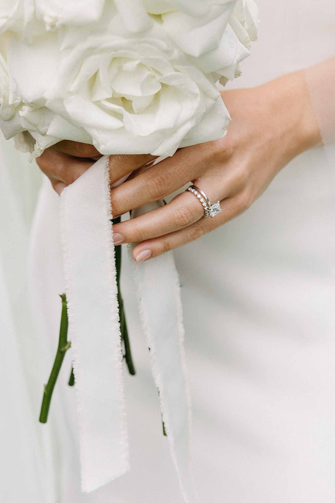 Close-up of bride's hand with diamond ring holding white rose bouquet wrapped in ivory silk ribbon