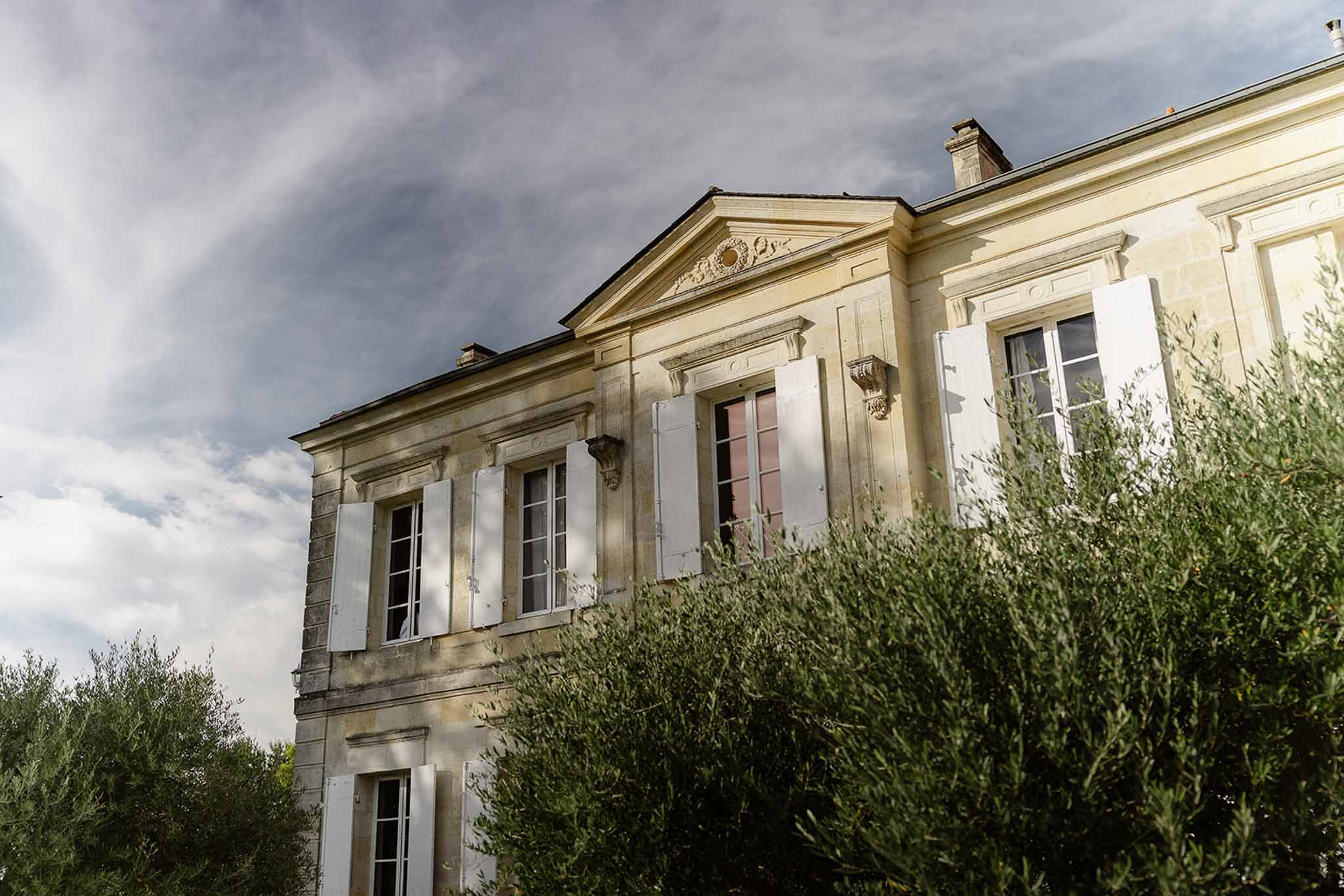 Upward angle of classical chateau facade with limestone masonry, white shutters, and olive trees
