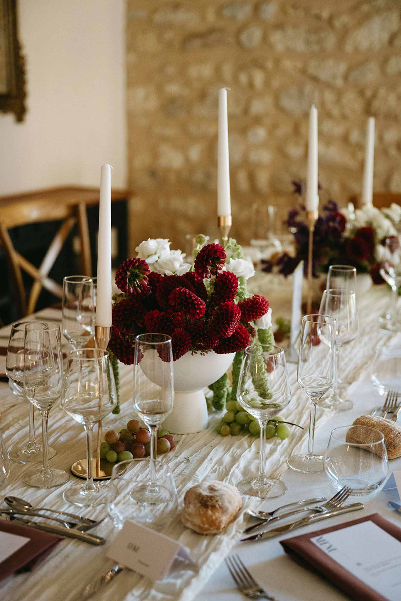 Crimson dahlia and ranunculus centerpiece with grapes, bread, and gold candlesticks on ivory runner