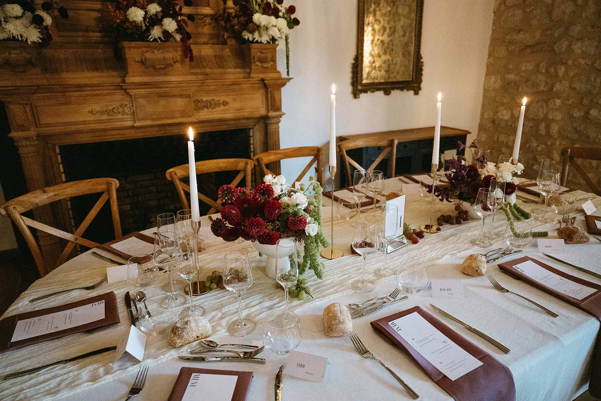 Reception table with crimson dahlias, white ranunculus, gold candlesticks, and mauve menus beside carved fireplace