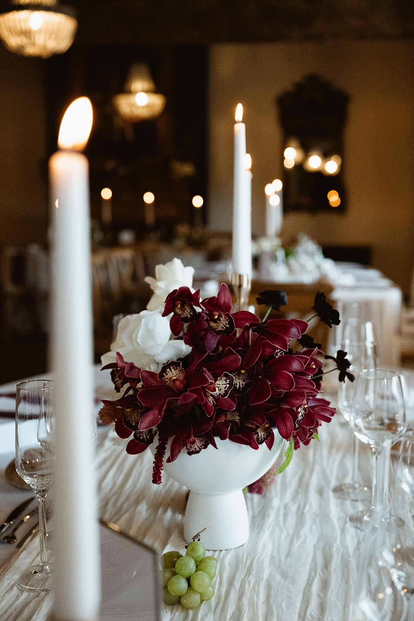 Candlelit reception table with burgundy orchid and white rose centerpiece, crystal glasses, and taper candles