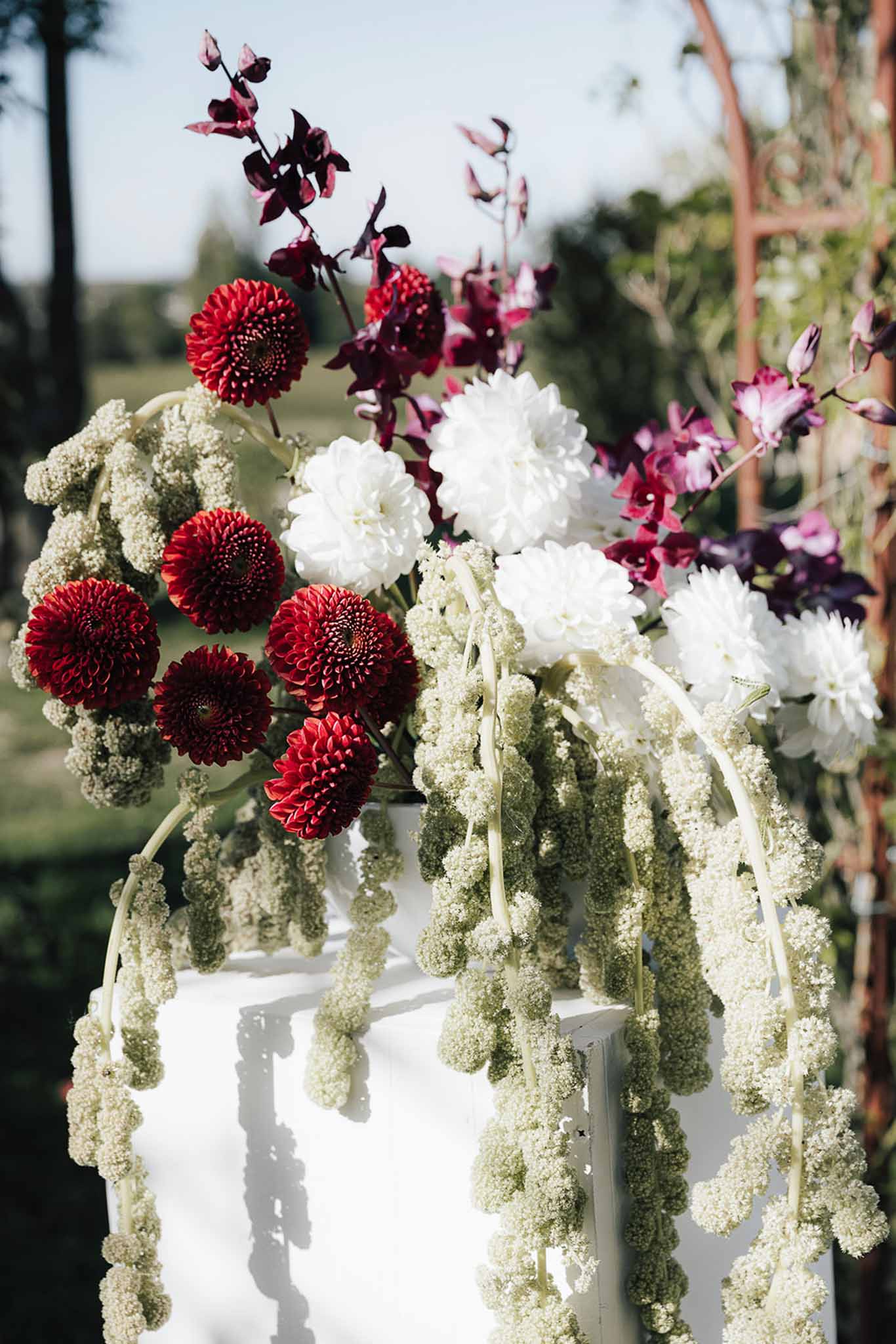 Ceremony floral arrangement on white pedestal with deep crimson dahlias, burgundy orchids, and trailing sage amaranthus