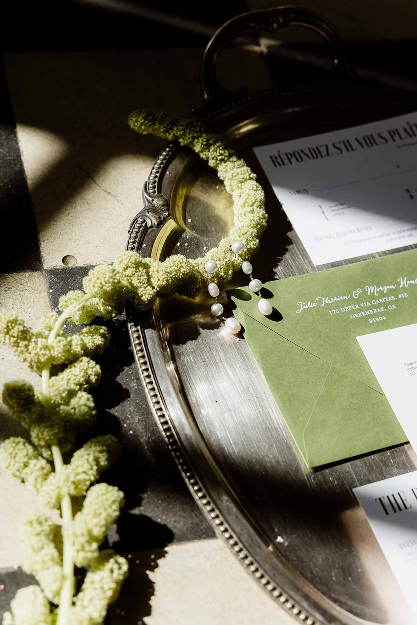 Flat lay of forest green envelope with white calligraphy, RSVP card, pearl earrings and greenery on silver tray