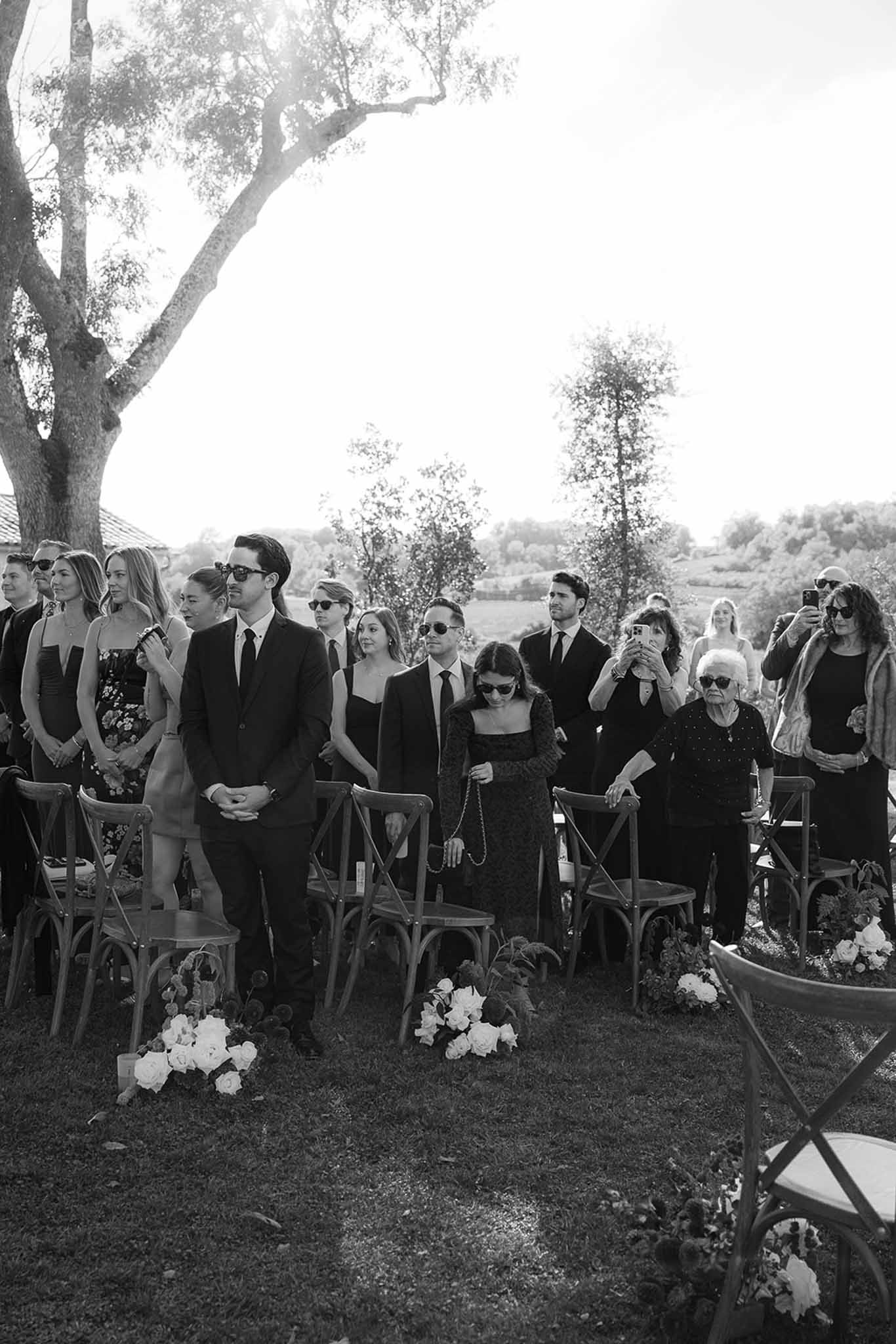 Outdoor wedding ceremony under large tree with guests in formal attire and white floral arrangements