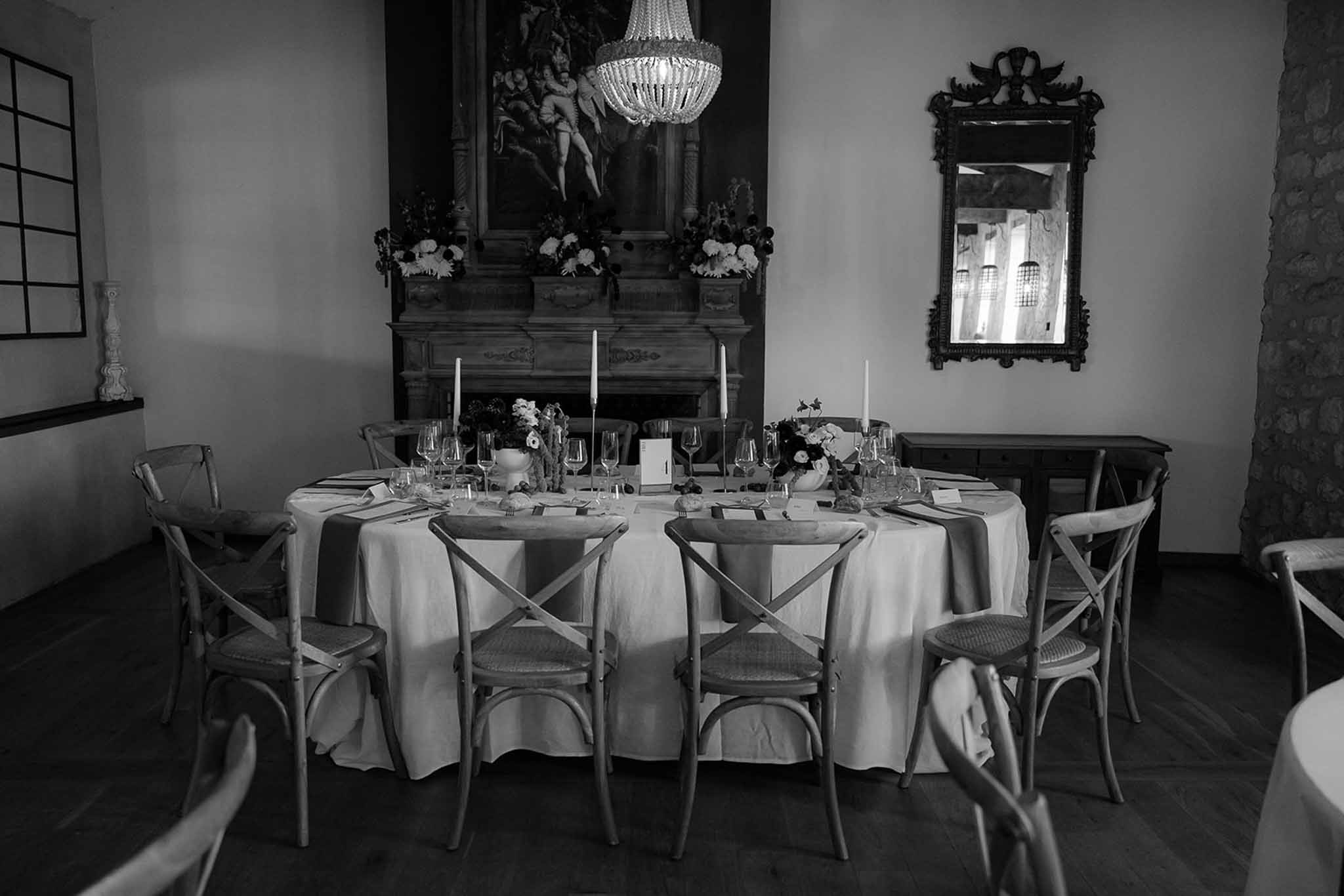 Black-and-white photo of round reception table with cross-back chairs, stone fireplace, chandelier, and oil painting