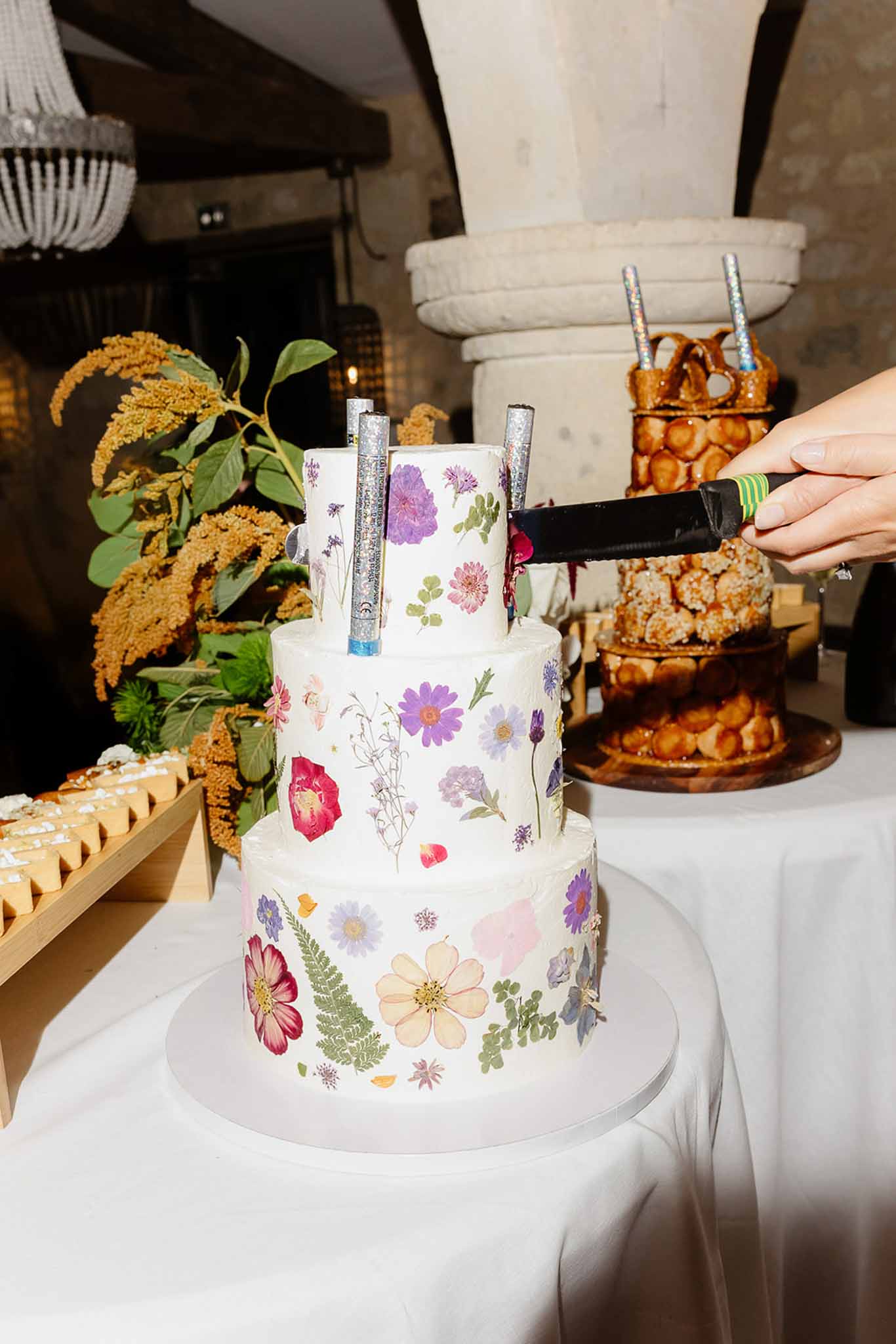 Three-tier pressed flower cake with sparklers beside croquembouche in stone-vaulted venue