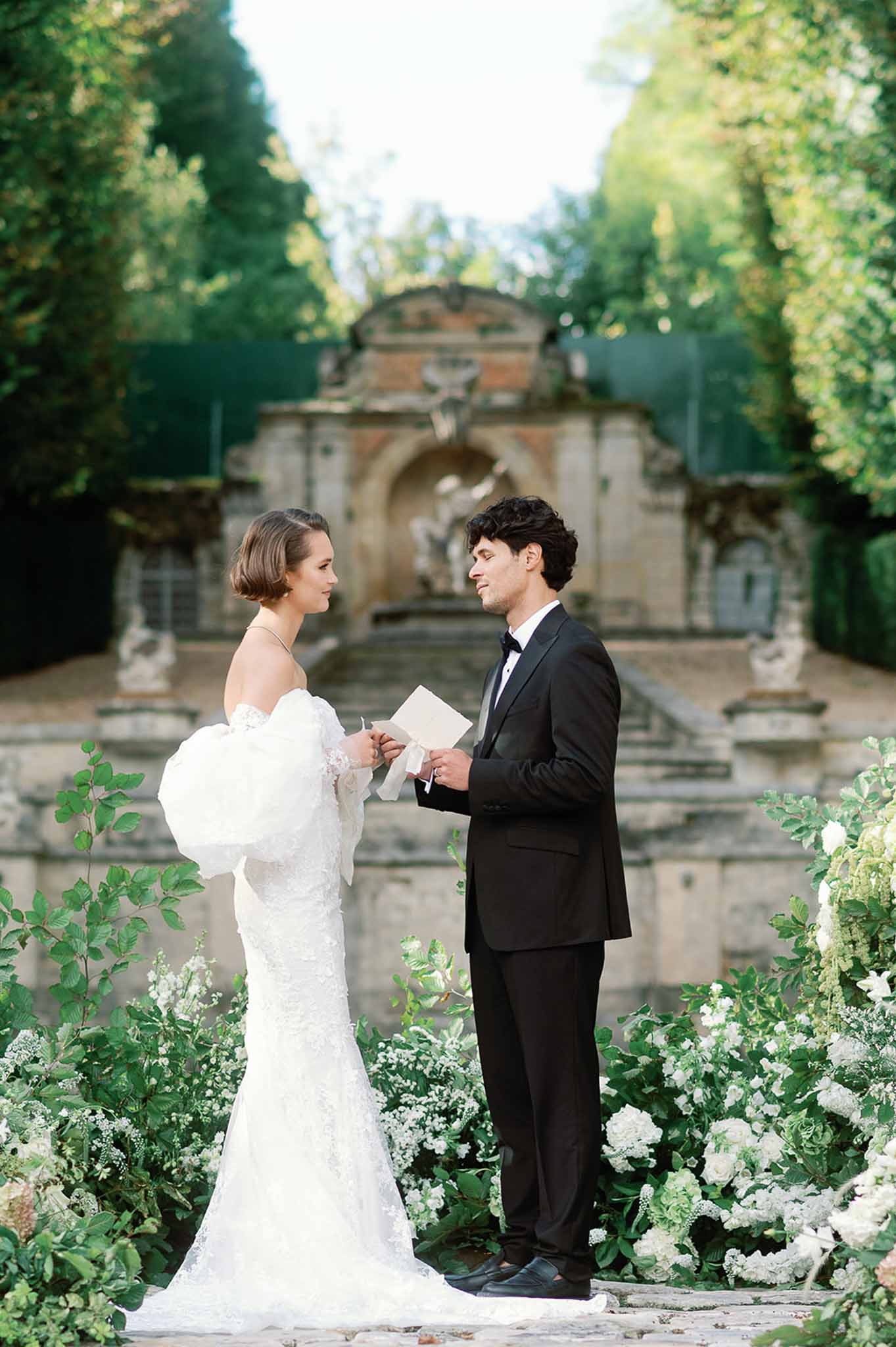 Bride and groom exchanging vows during outdoor ceremony in formal garden with hedge walls and stone architecture