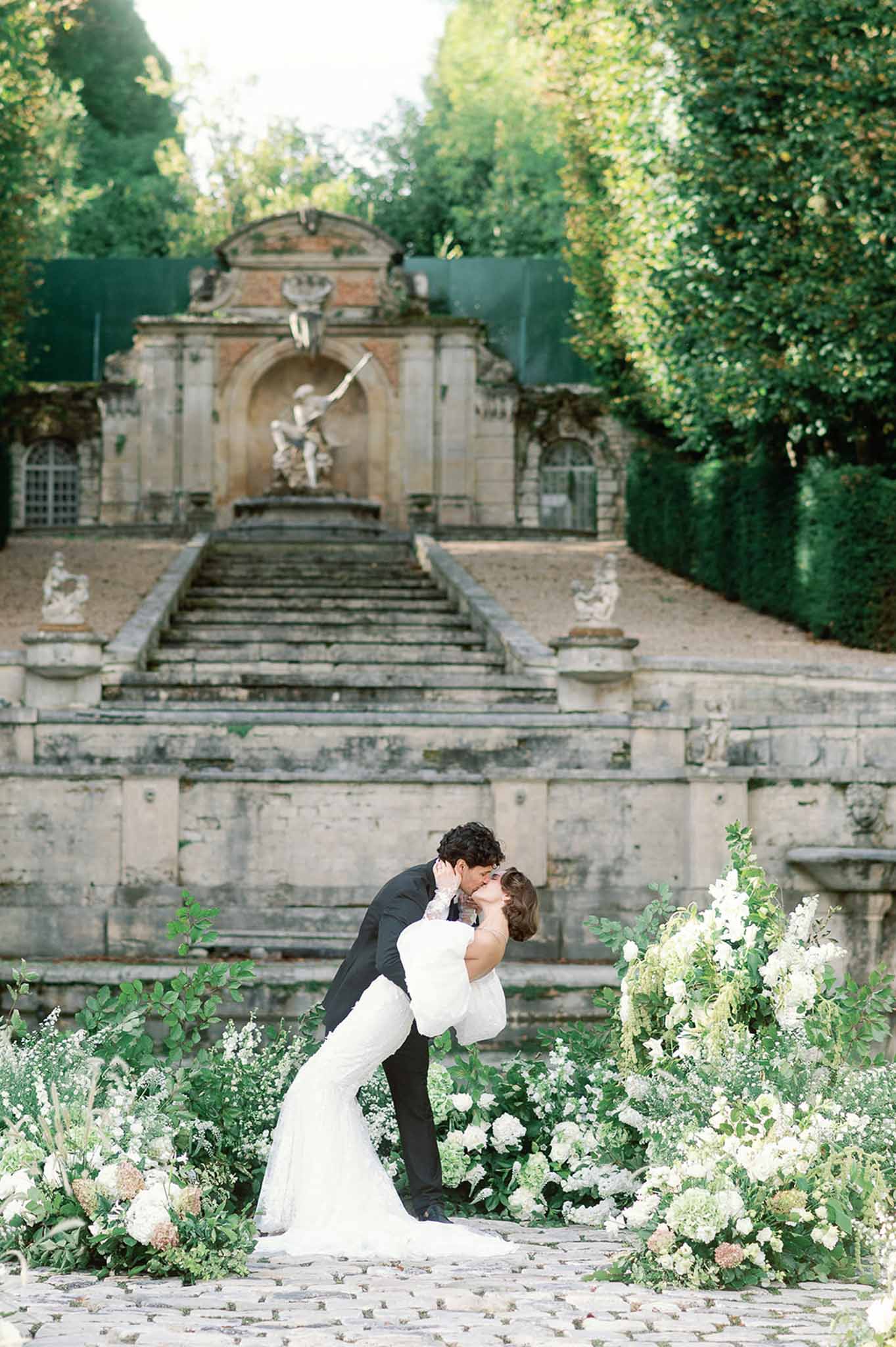 Bride and groom kissing in romantic dip pose surrounded by white roses in formal garden courtyard