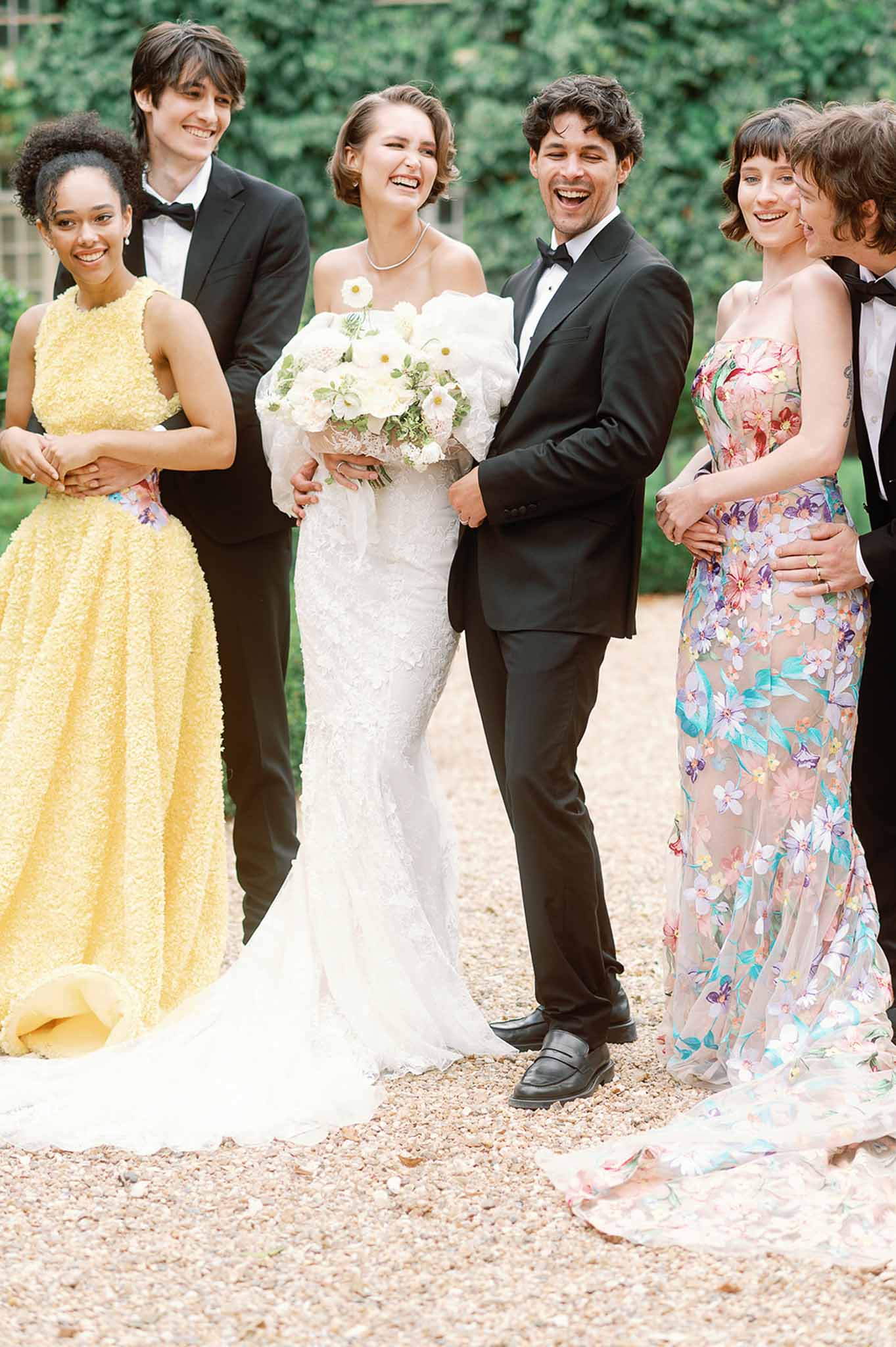 Bridal party portrait with bride, groom, and wedding party on outdoor gravel pathway with ivy-covered walls