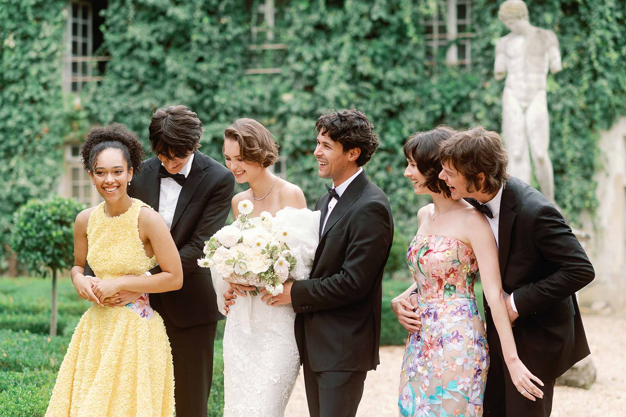 Wedding party walking together in formal garden with ivy-covered walls and manicured lawn