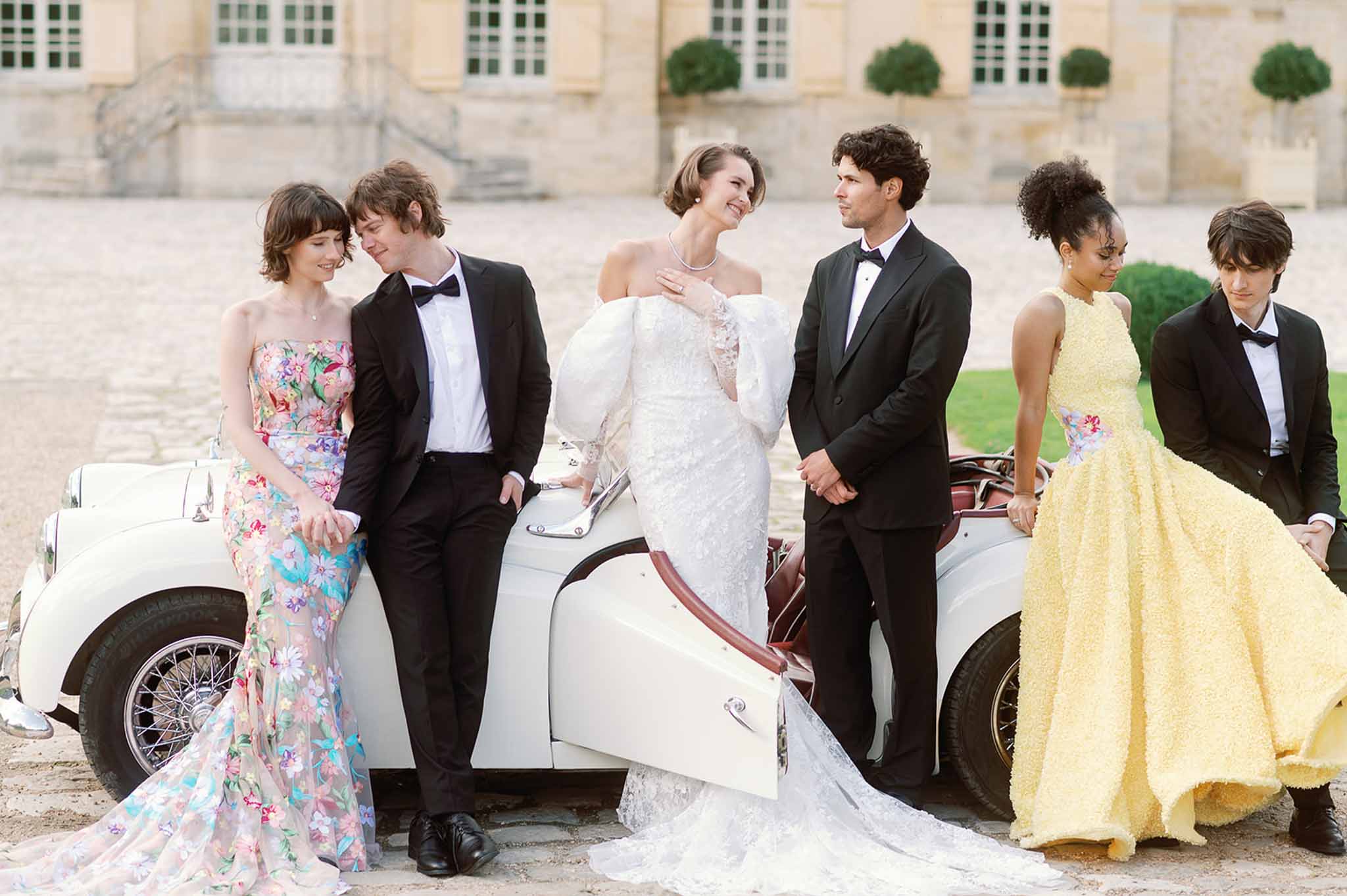 Wedding party posing with vintage convertible cars in stone château courtyard
