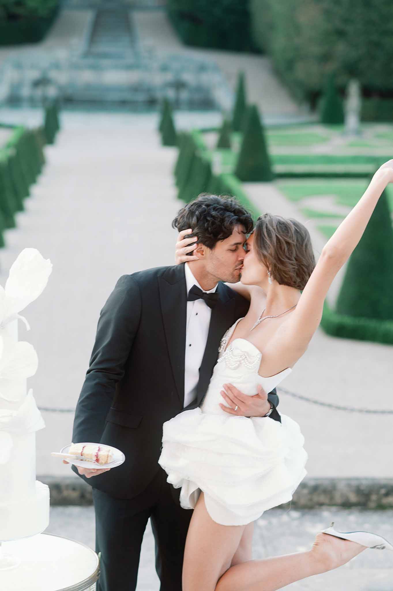 Bride and groom sharing kiss during cake cutting at formal garden reception