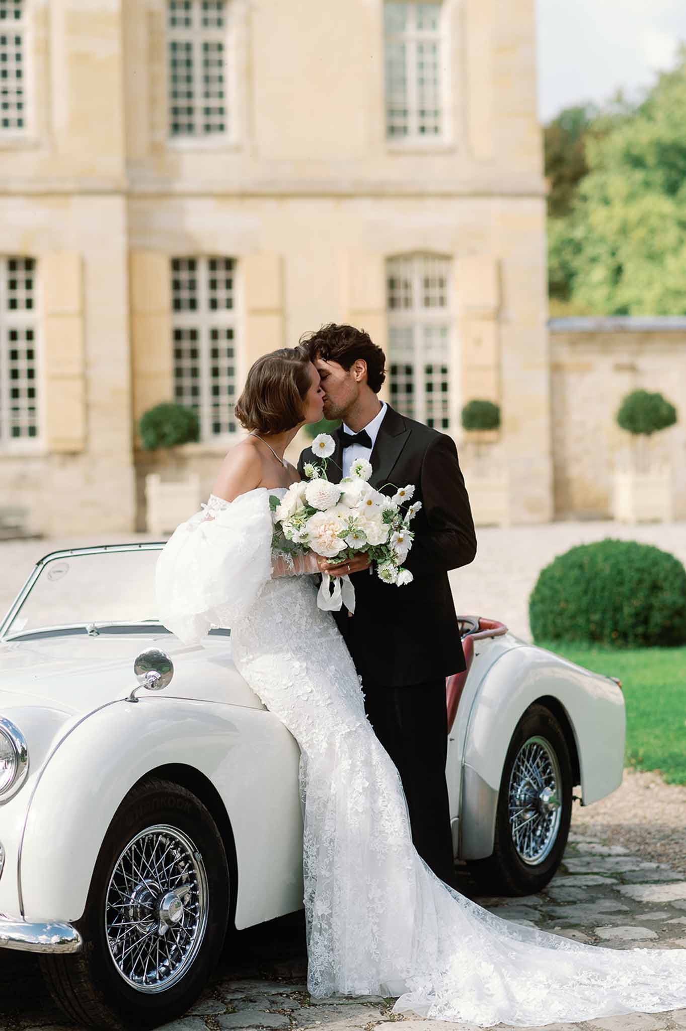 Bride and groom kissing beside vintage convertible in French château courtyard