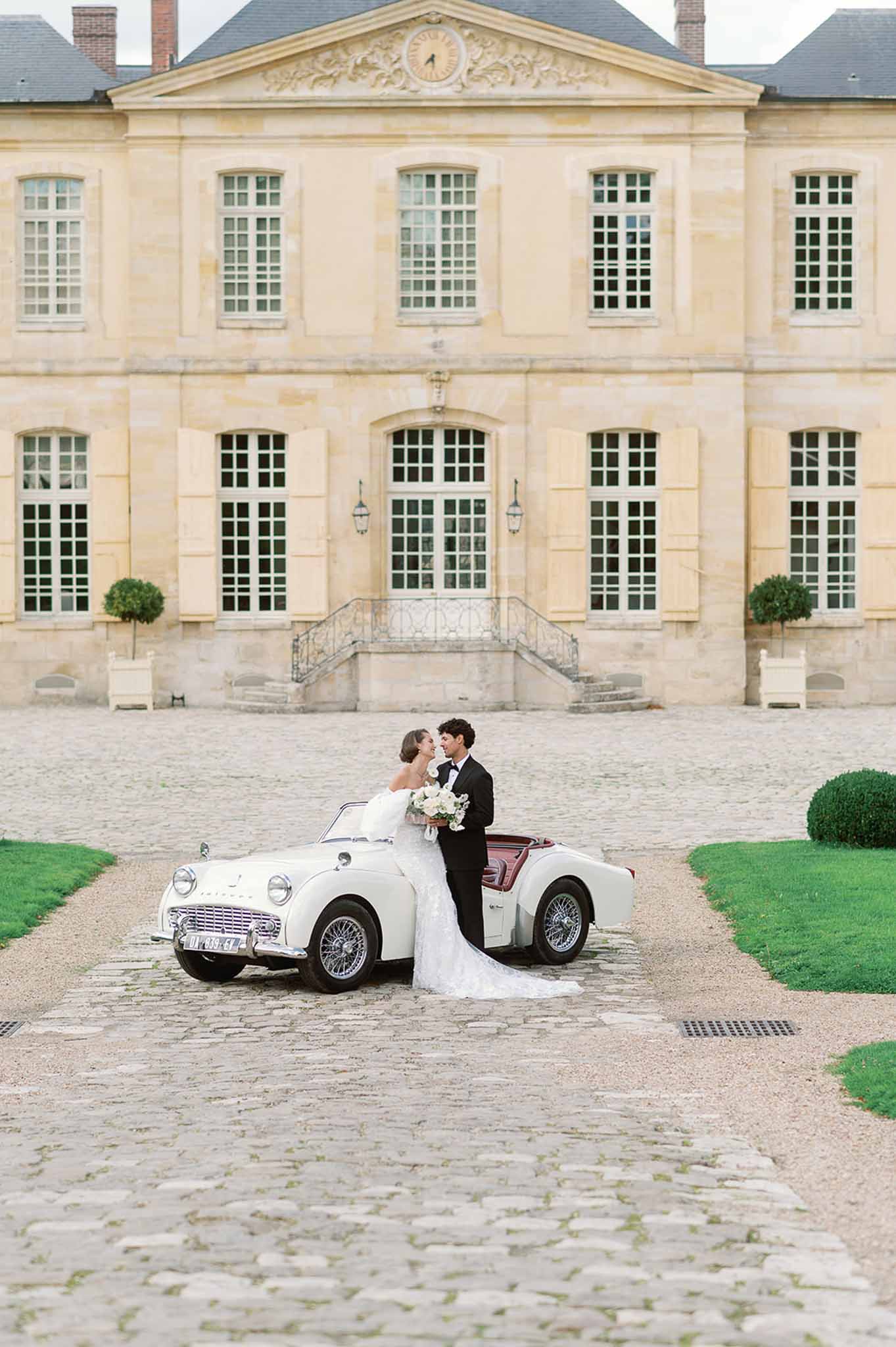 Bride and groom with vintage convertible car at classical stone château courtyard