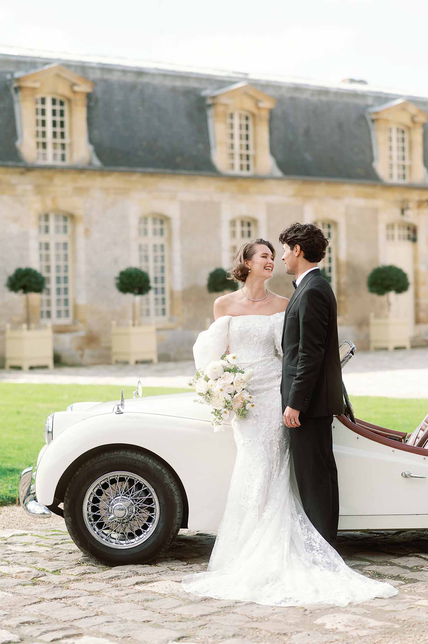 Newlyweds posing beside vintage white convertible at French château courtyard