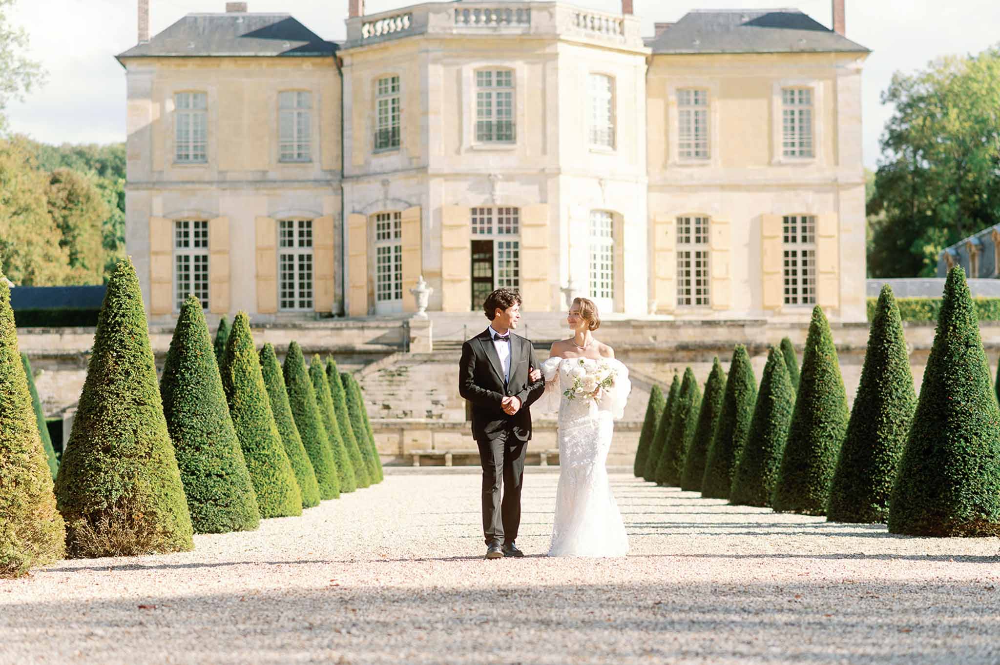 Bride and groom walking through formal topiary garden at neoclassical château
