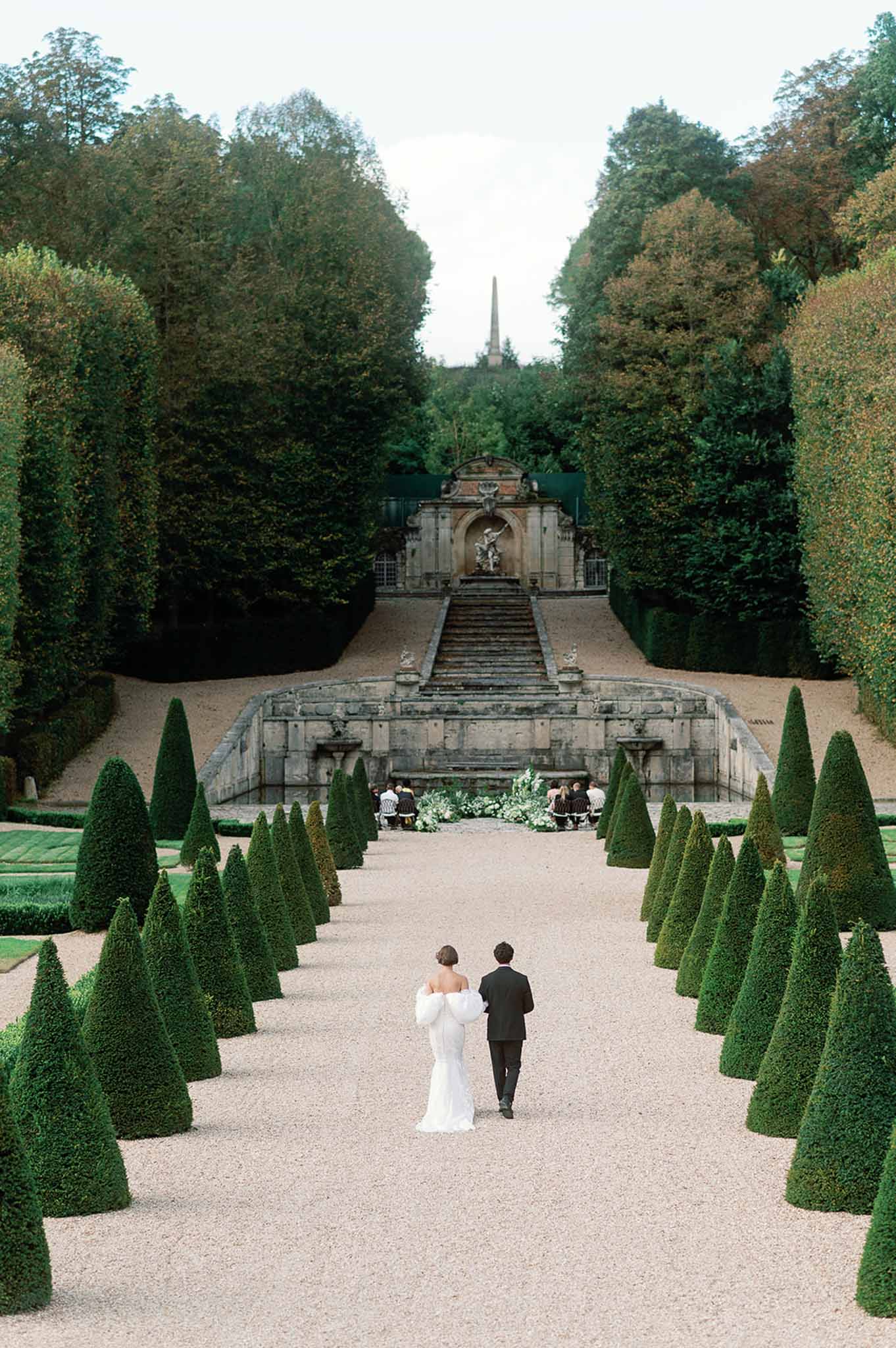 Bride and groom walking down formal garden pathway at French estate with topiary and classical architecture