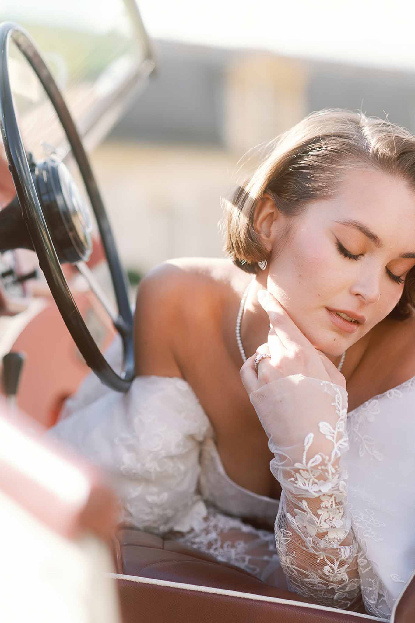 Bride in ivory lace dress sitting in vintage car adjusting veil during wedding portrait session