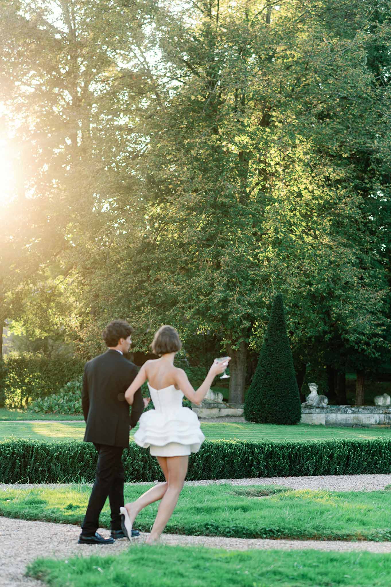 Bride and groom walking together through formal garden with manicured hedges and cypress trees