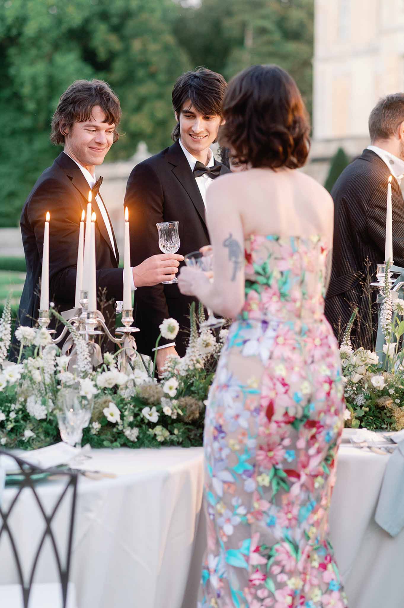 Cocktail hour guests conversing on outdoor terrace with candlelit table settings and floral arrangements
