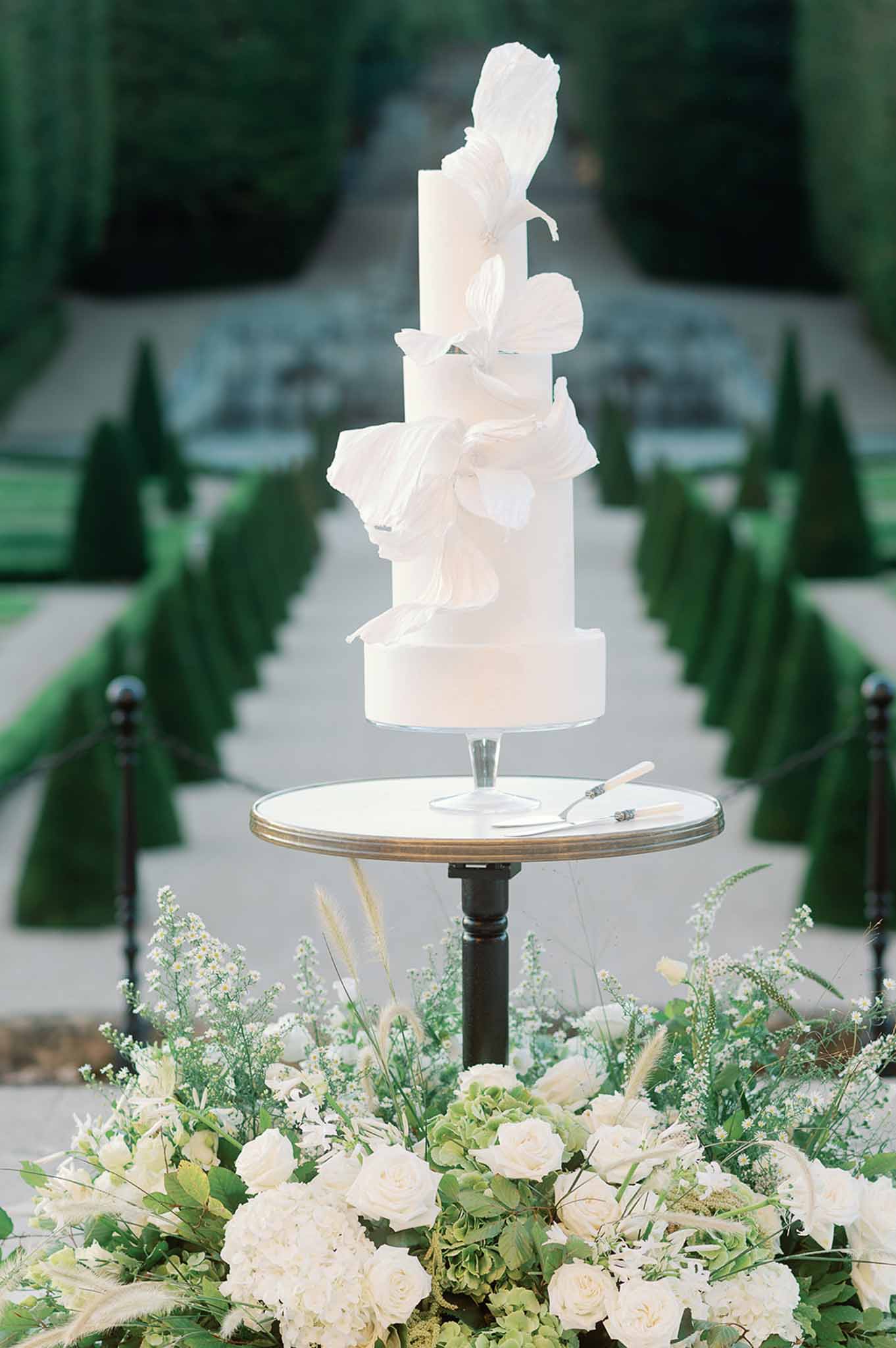 Four-tiered white wedding cake with sugar flowers displayed on brass-trimmed table in formal garden setting