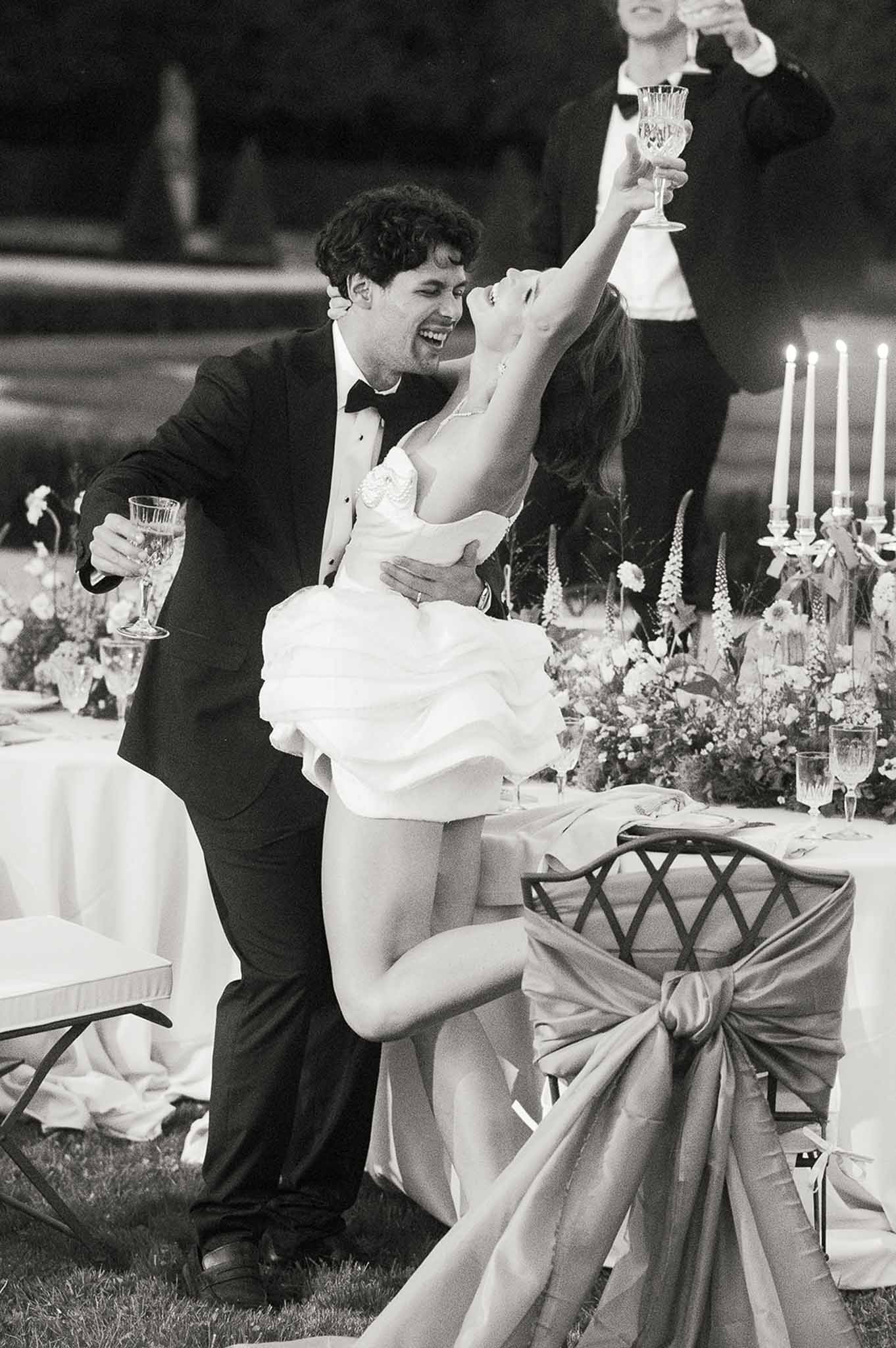 Groom lifting bride with champagne flute during outdoor evening reception celebration