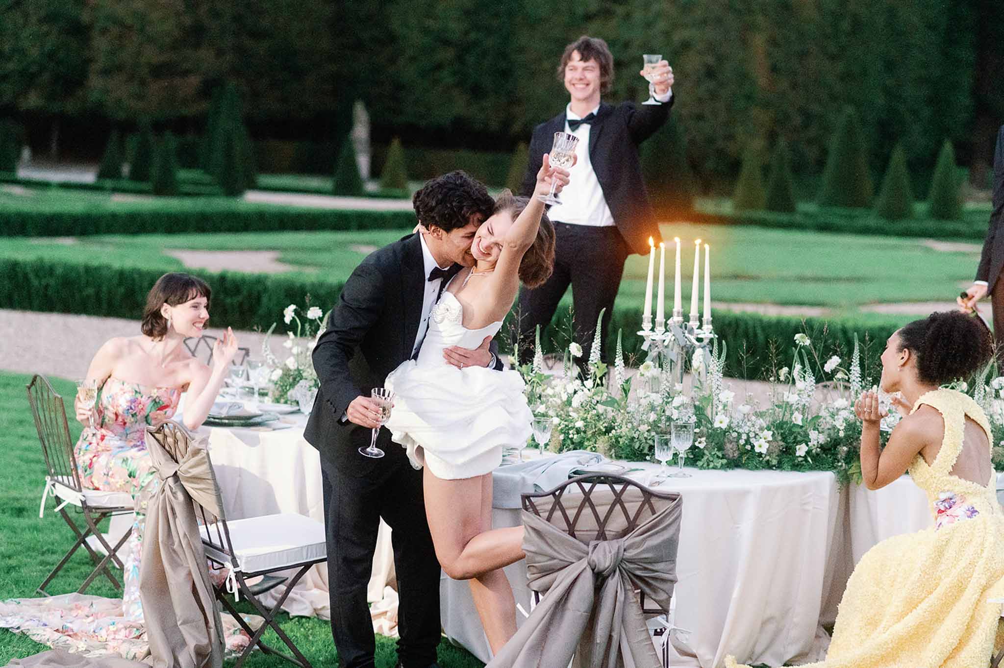 Groom lifting bride during reception dinner toast on manicured garden lawn with formal table setting