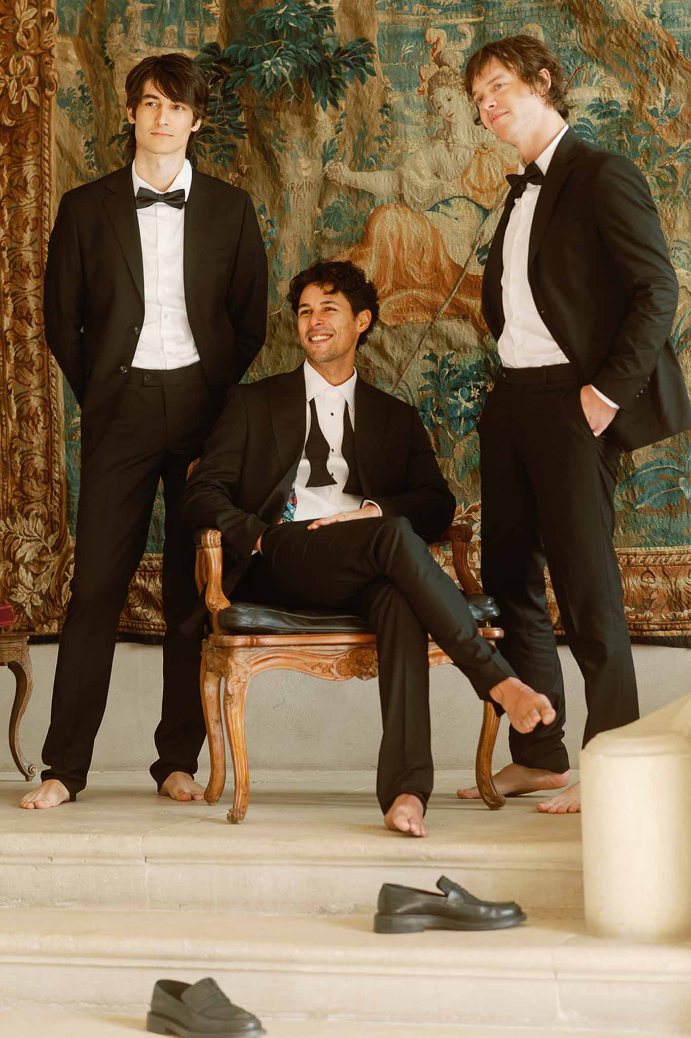 Four groomsmen in black tuxedos posing indoors against ornate tapestry backdrop