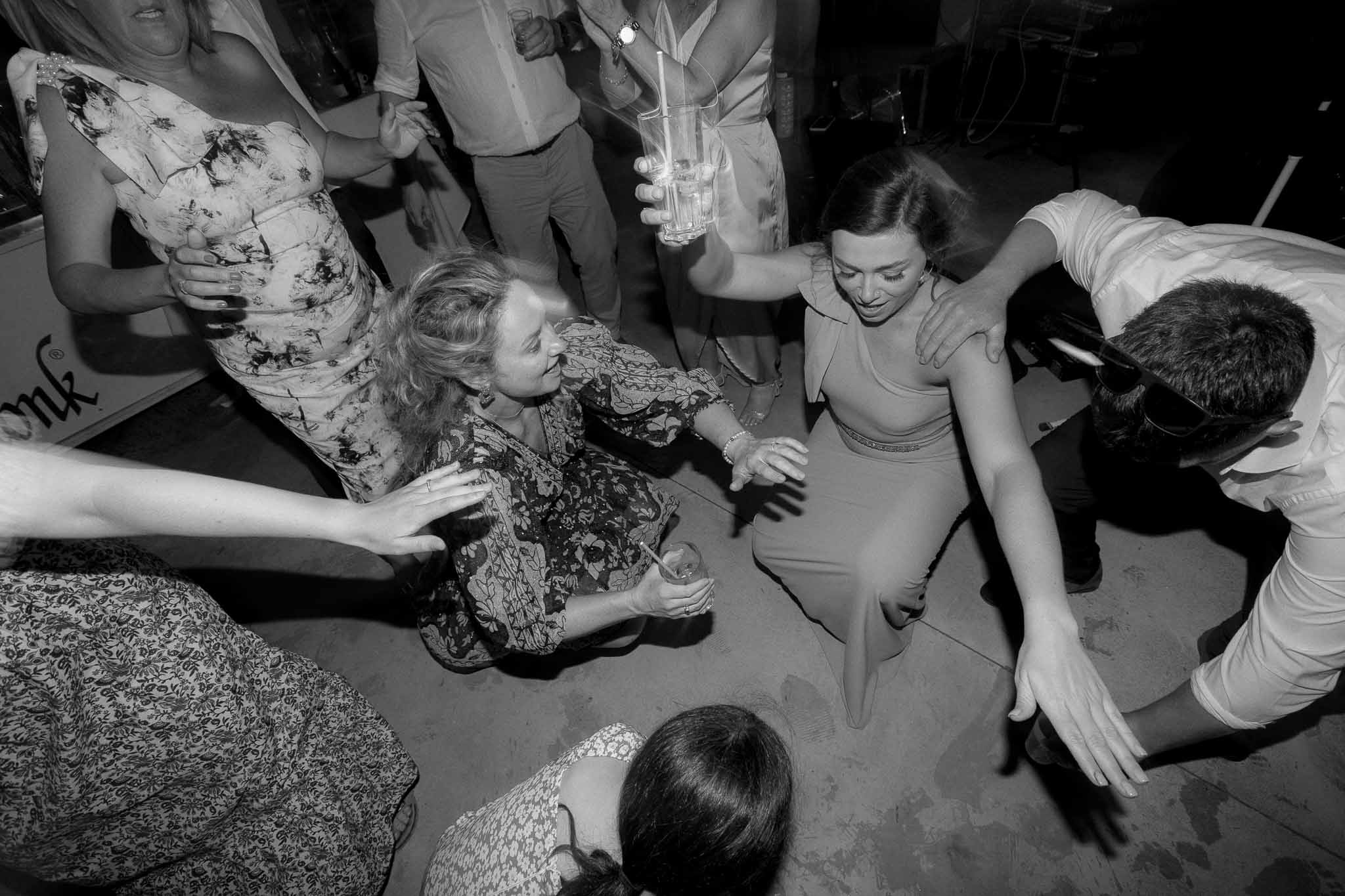 Black and white high-energy dance floor shot of guests crouching and leaning in with drinks