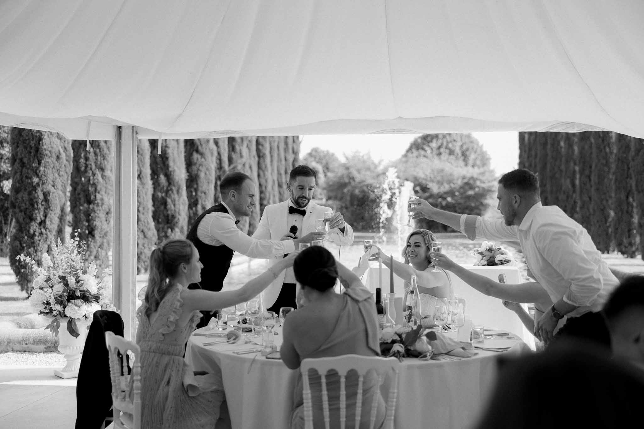 Groom in white dinner jacket leading toast at round reception table under marquee in black and white