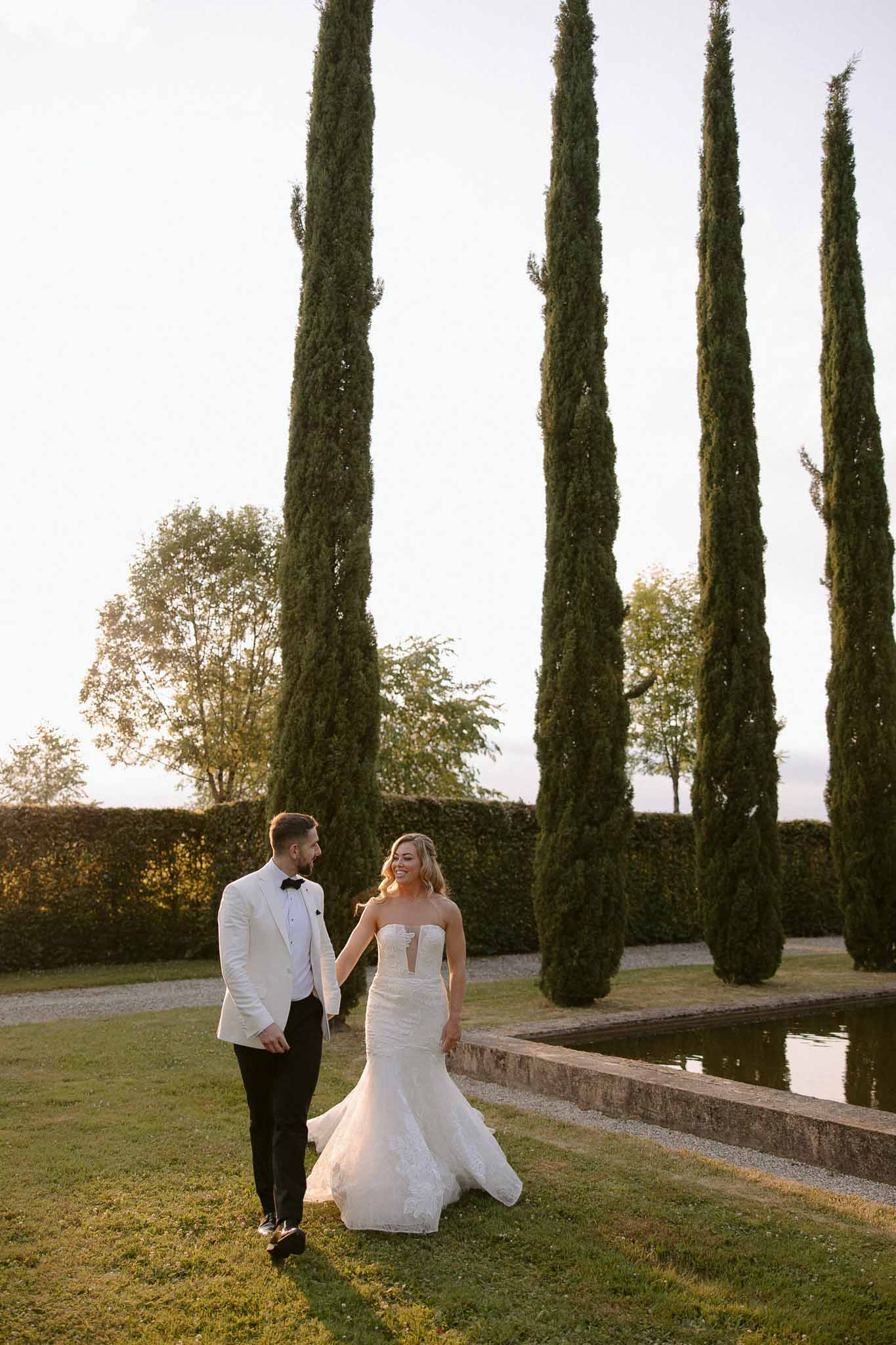 Bride and groom walking hand-in-hand through formal garden with reflecting pool and cypress trees