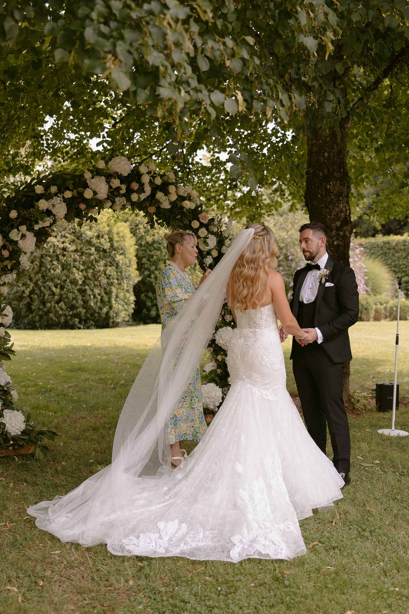 Bride and groom during outdoor garden ceremony with floral arch and attendant adjusting veil