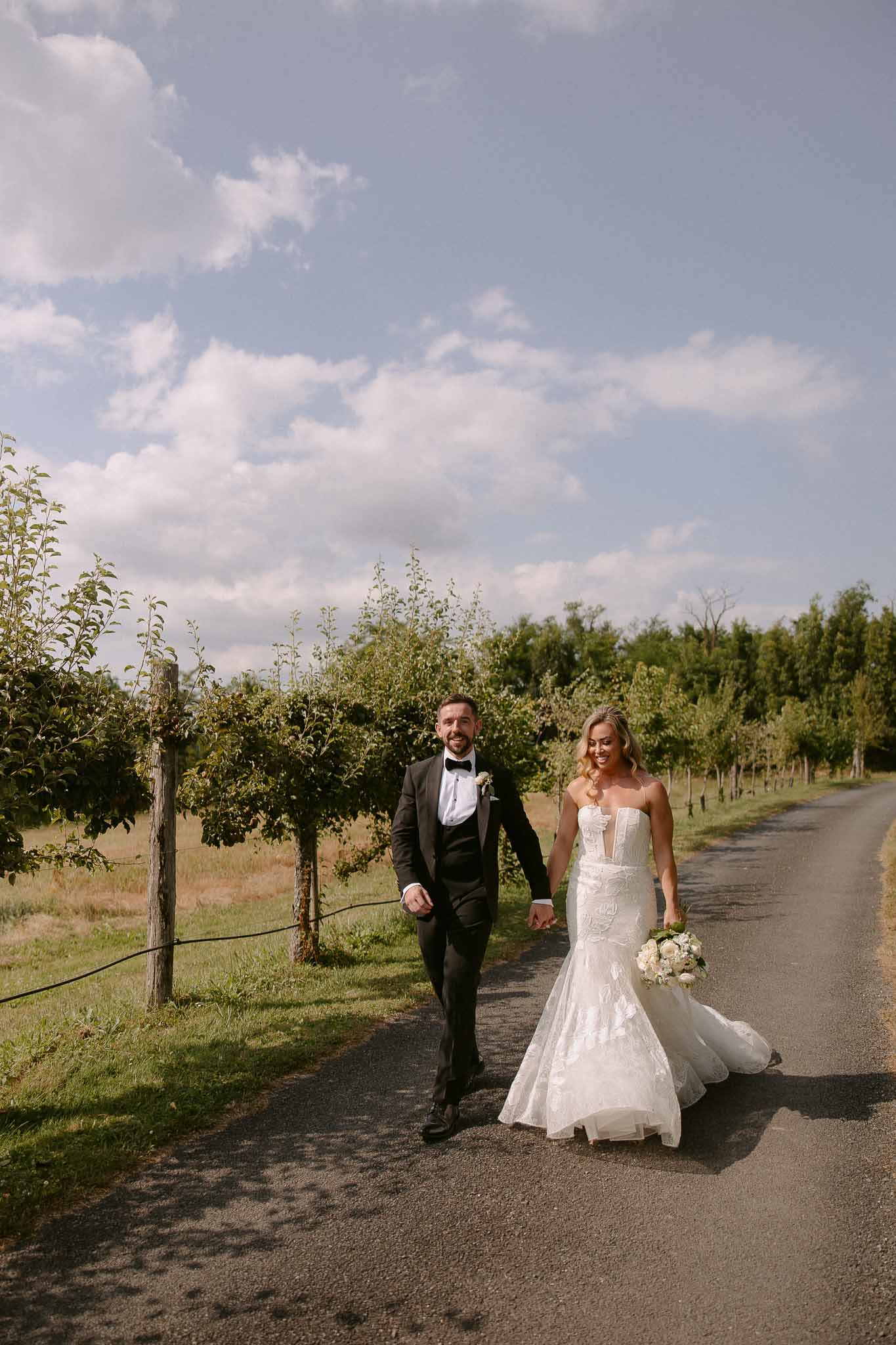 Bride and groom walking hand-in-hand down tree-lined vineyard road after wedding ceremony