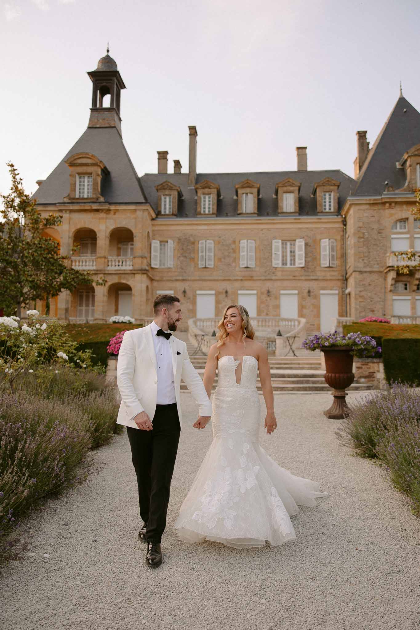 Bride and groom walking hand-in-hand along château courtyard pathway with lavender hedges and stone architecture