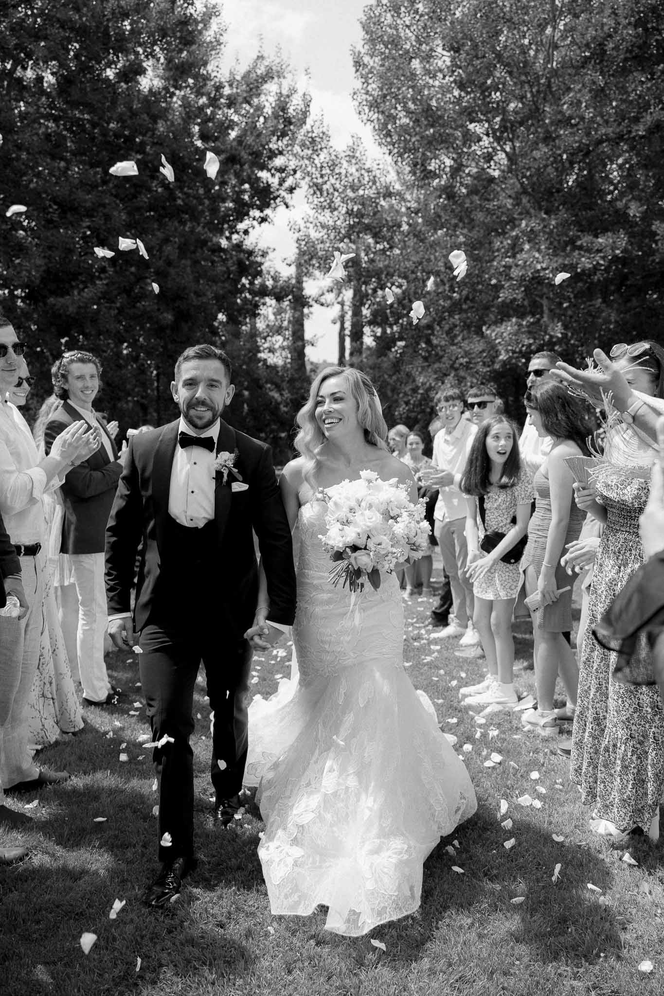 Newlyweds walking down cypress-lined aisle with guests throwing confetti at outdoor wedding ceremony