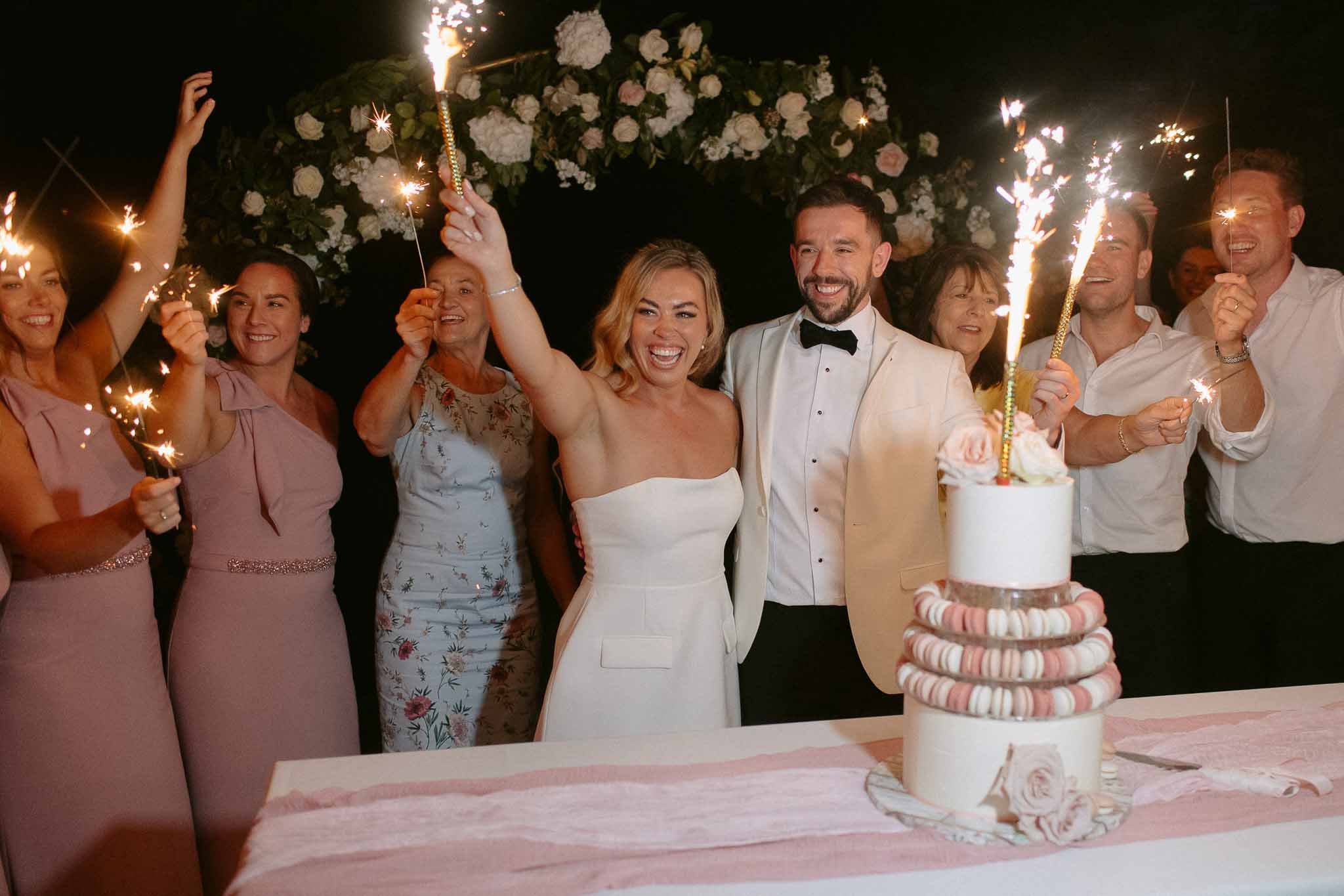 Couple with sparklers at macaron wedding cake beside white peony and blush rose arch at night