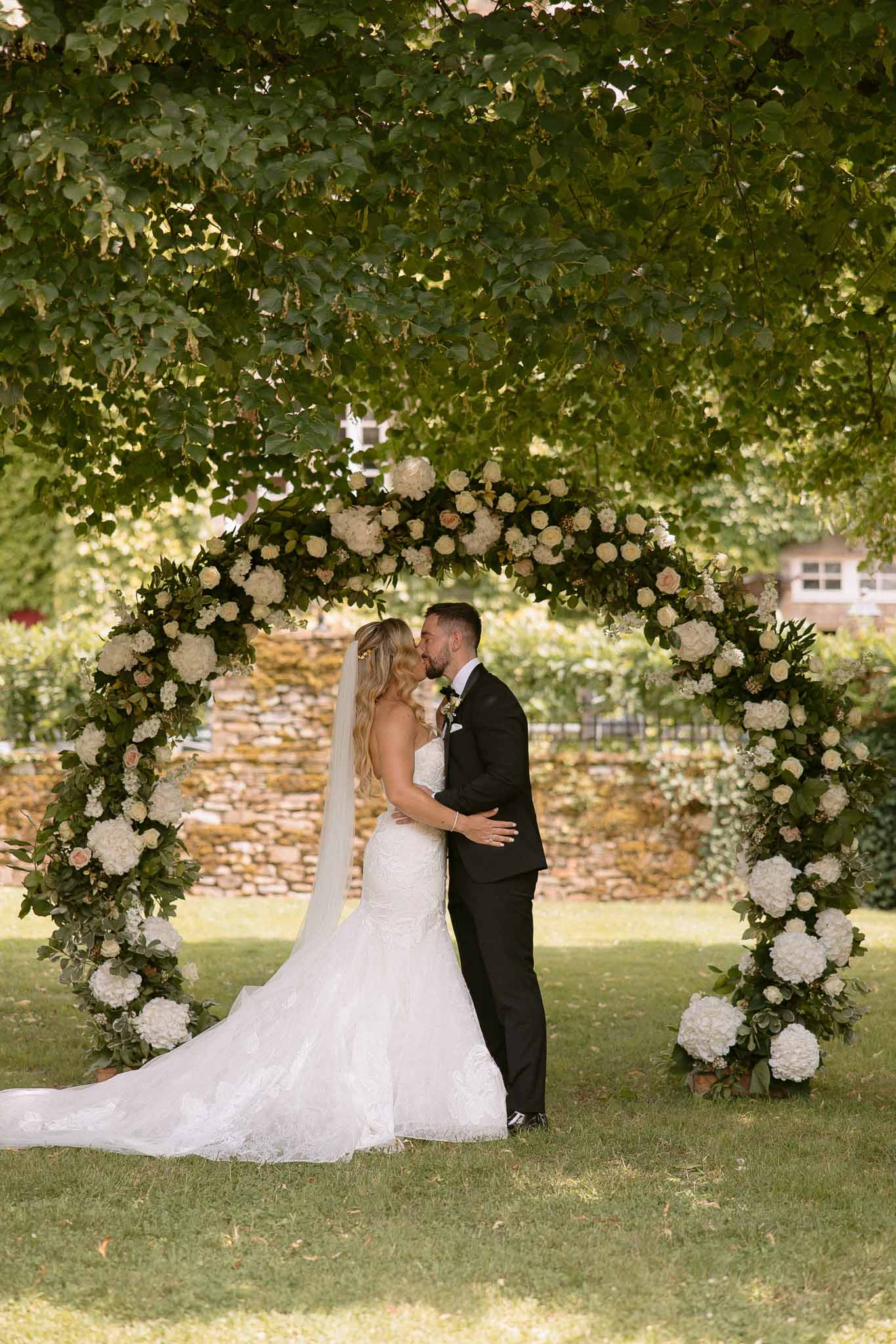 Bride and groom kissing under floral arch during outdoor garden ceremony at estate venue