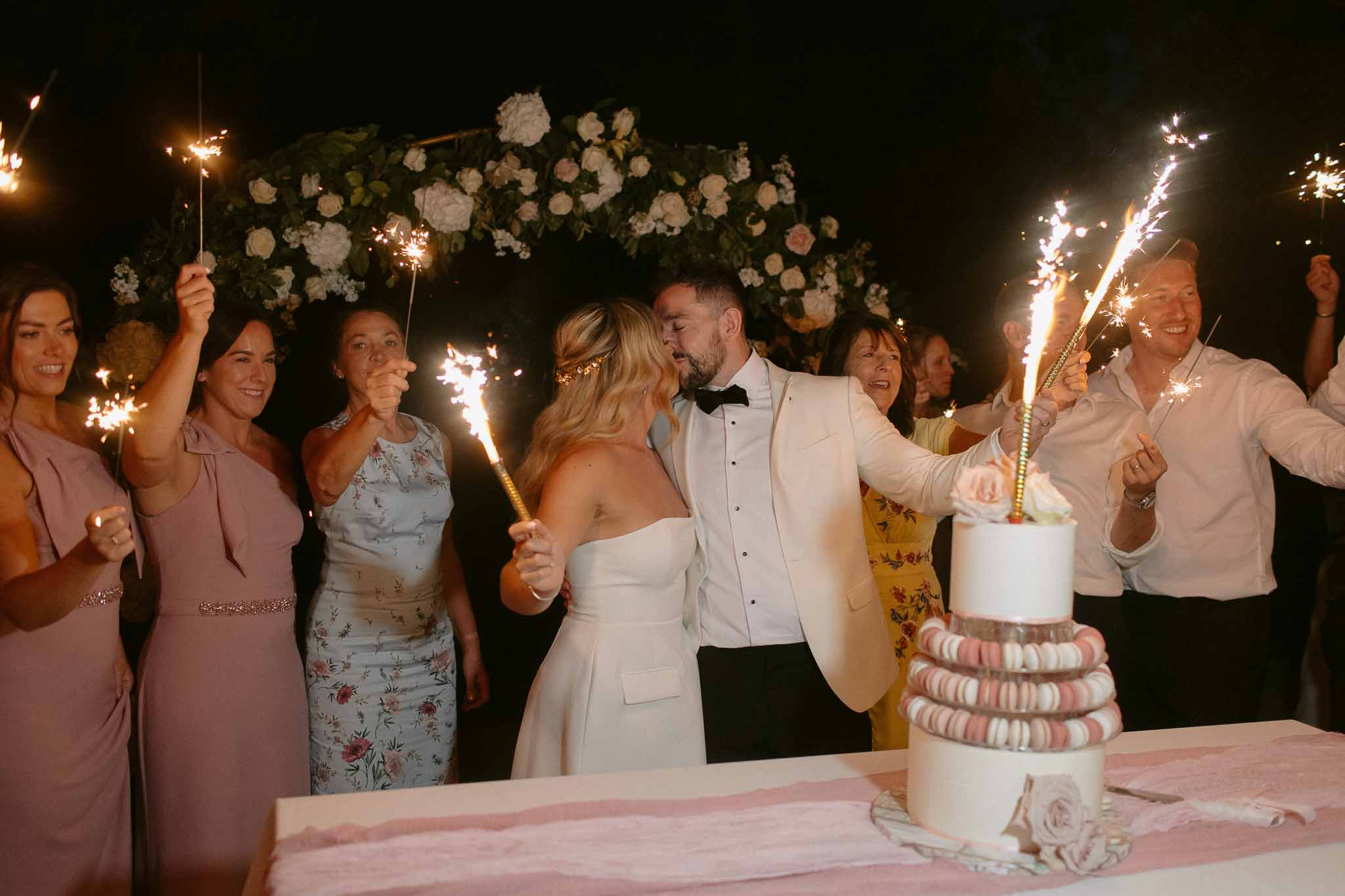 Bride and groom kiss at macaron cake with sparklers and floral arch at outdoor evening reception