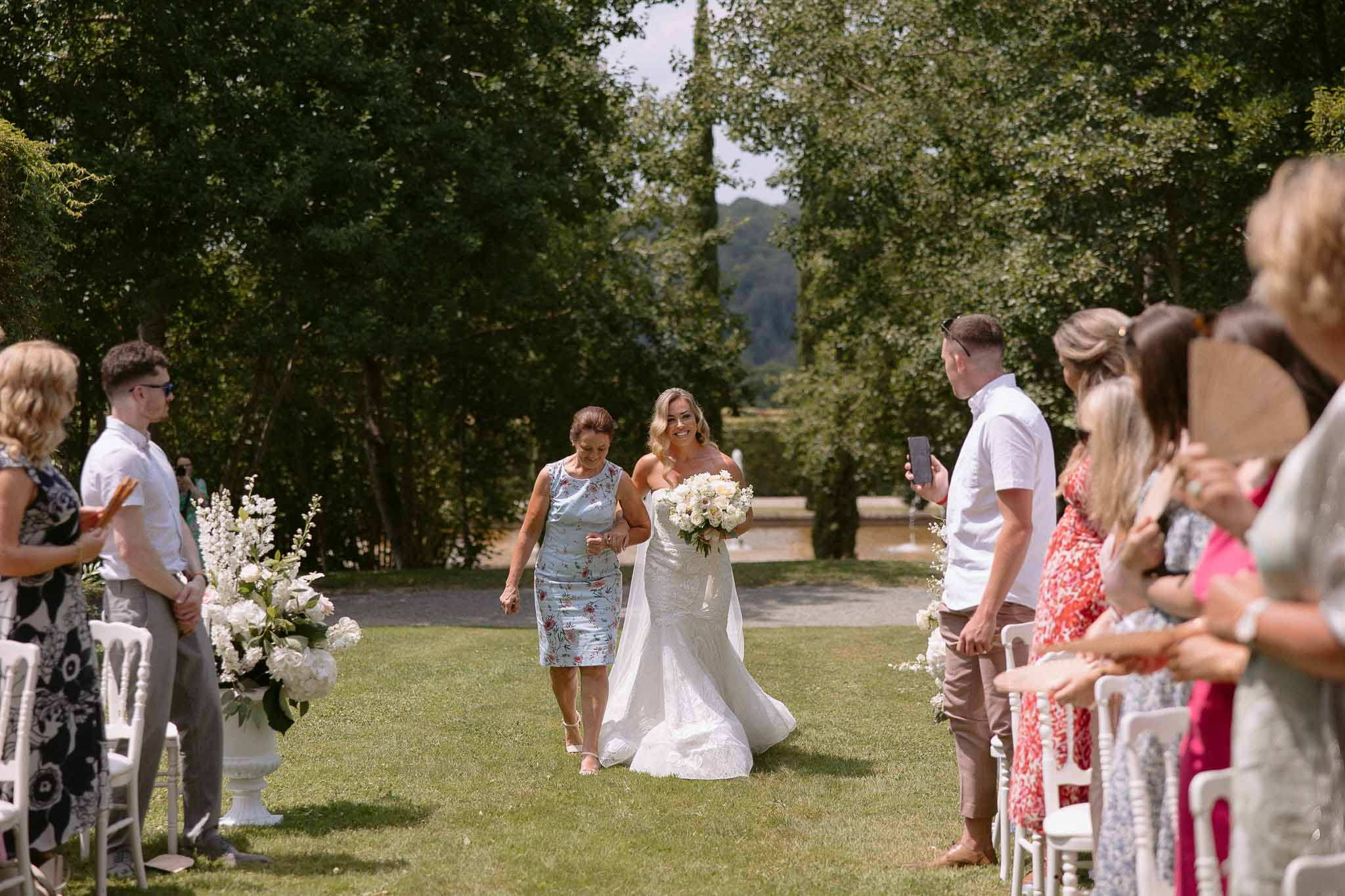 Bride walking down garden aisle during outdoor wedding ceremony with guests in white chairs