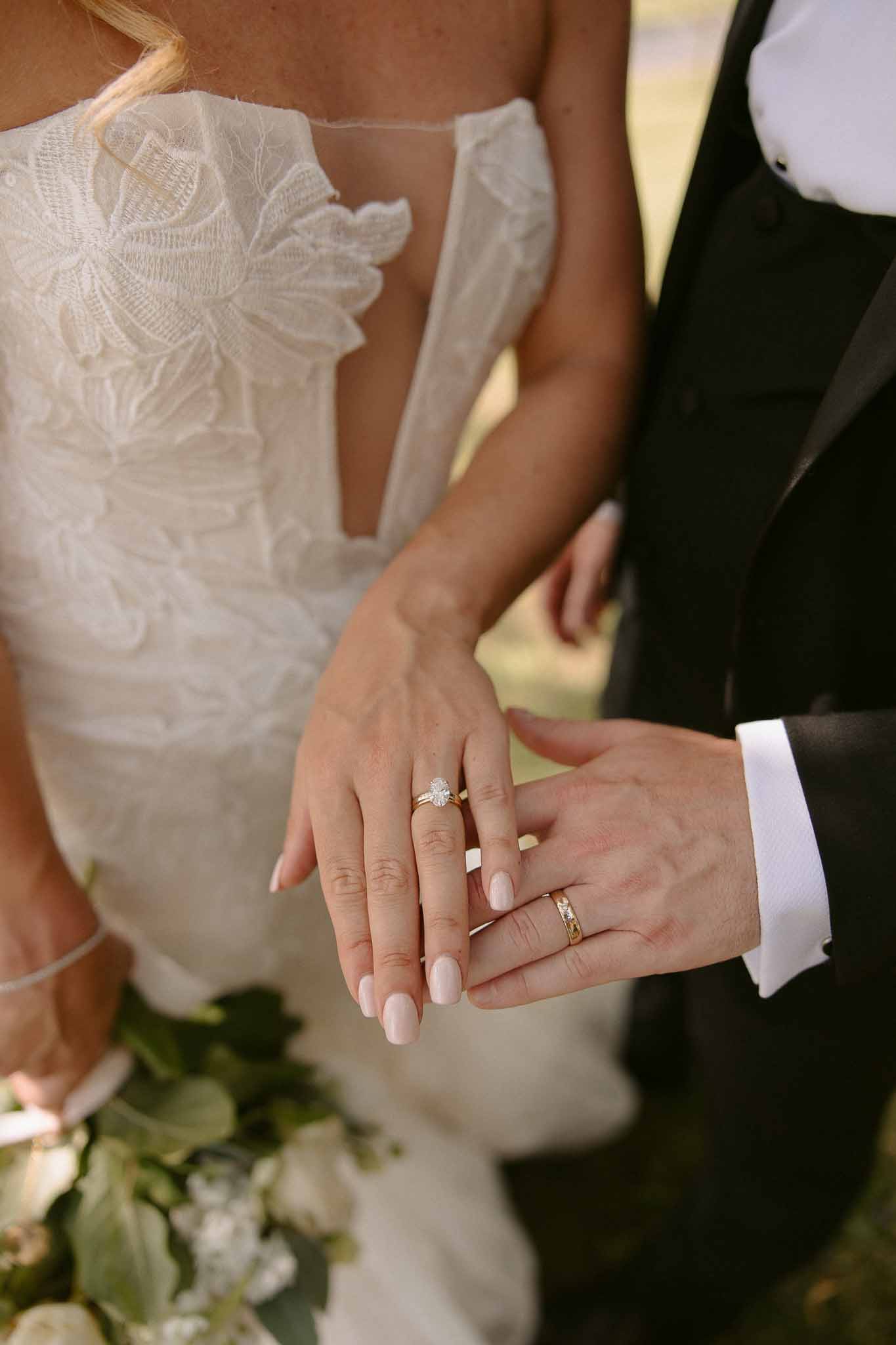 Close-up of bride and groom's hands showing gold wedding rings against white lace gown and black tuxedo