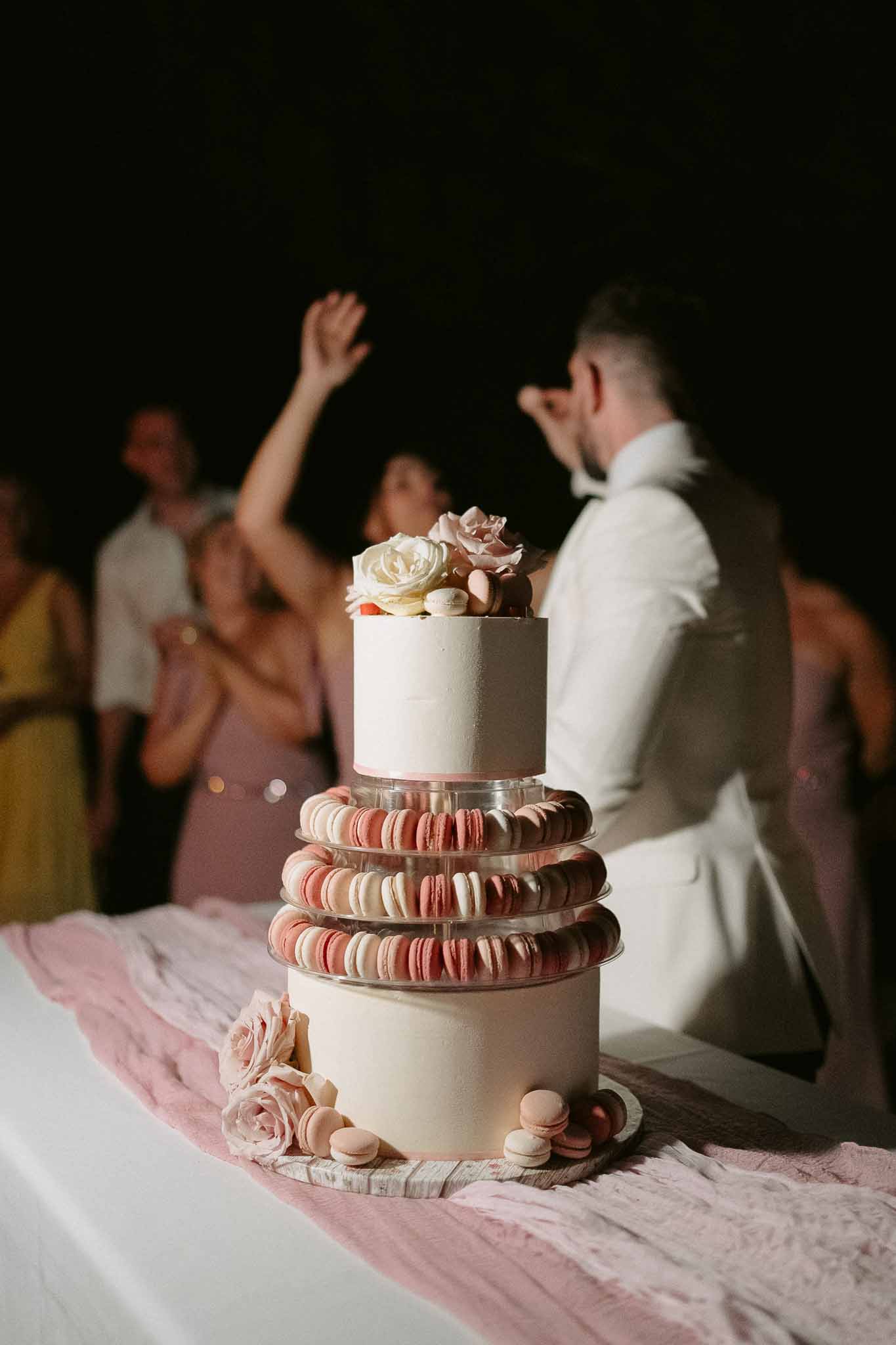 Two-tier ivory wedding cake with pink and white macaron layers, blush roses, and guests dancing in background